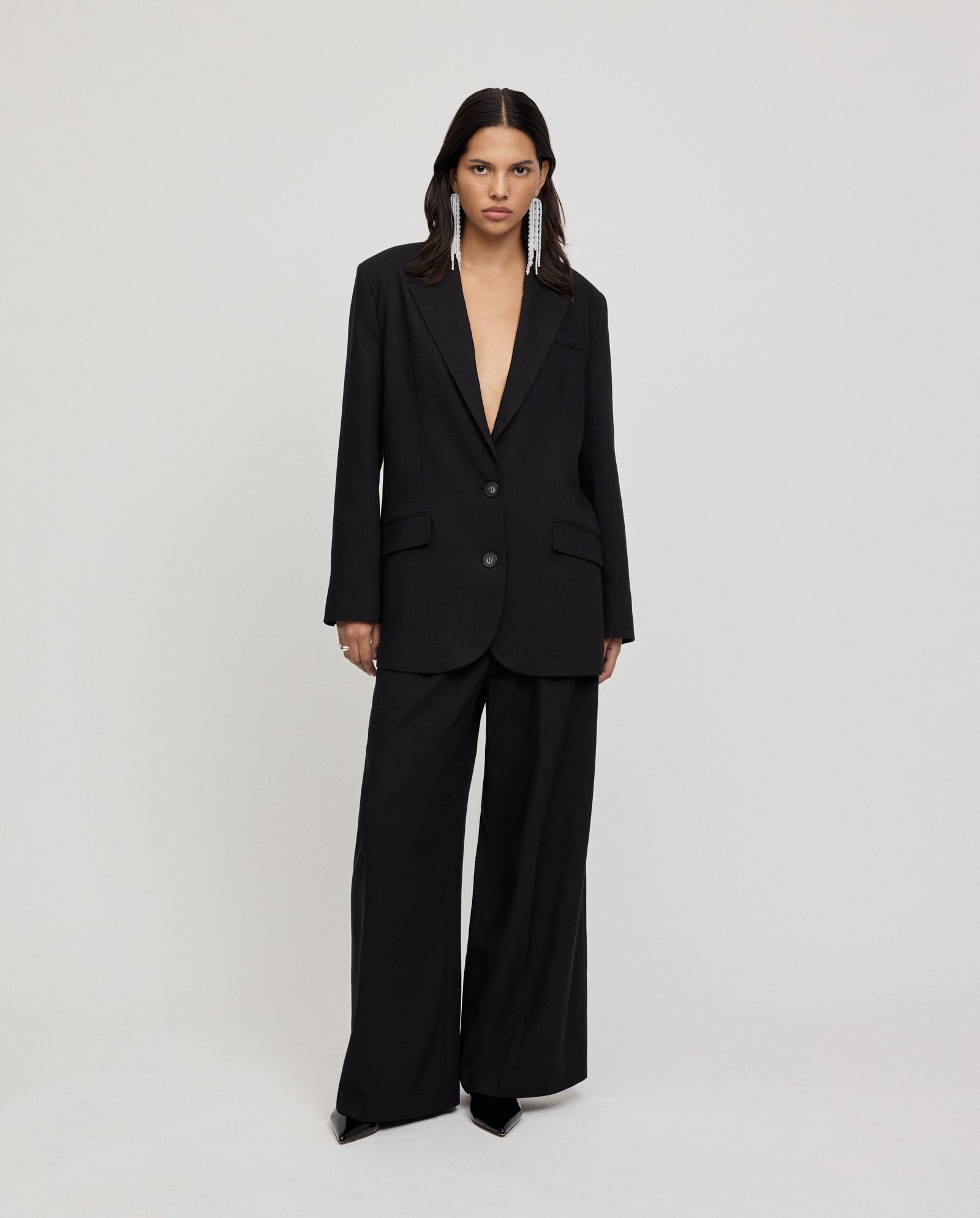 A woman stands against a plain background wearing IVY OAKs oversized black JUNA Blazer with a deep V and wide-leg pants, styled with long earrings.