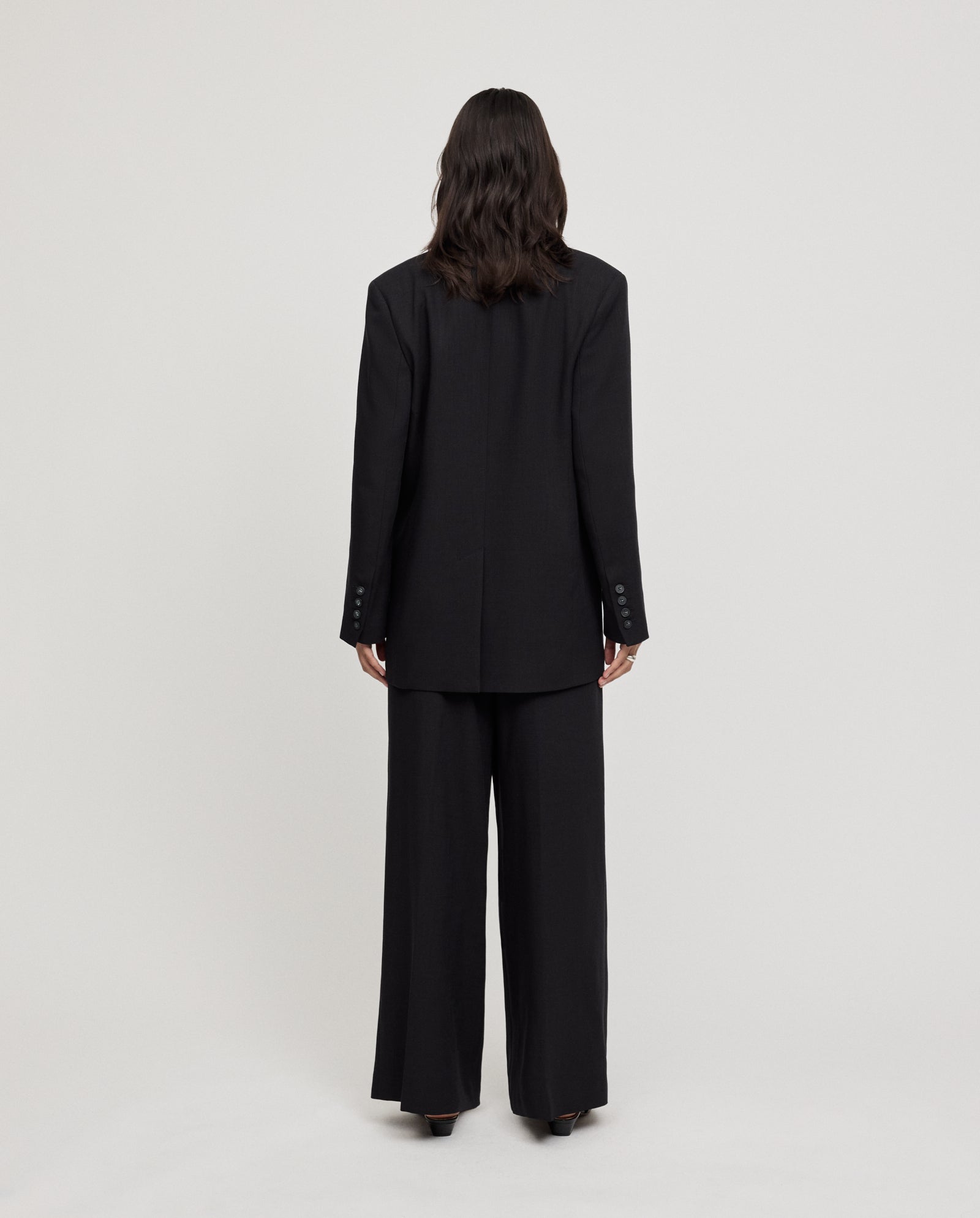 A person with wavy brown hair stands facing away against a plain background, wearing the IVY OAK JUNA Blazer—an oversized, single-breasted black piece.