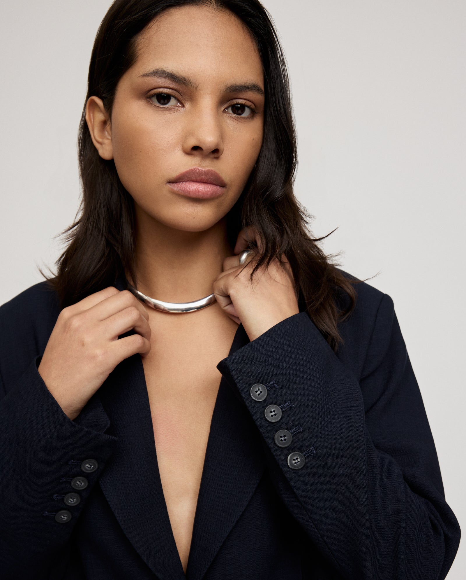 A woman confidently poses against a plain background, wearing a dark single-breasted JUNA Blazer from IVY OAK and holding a silver necklace.