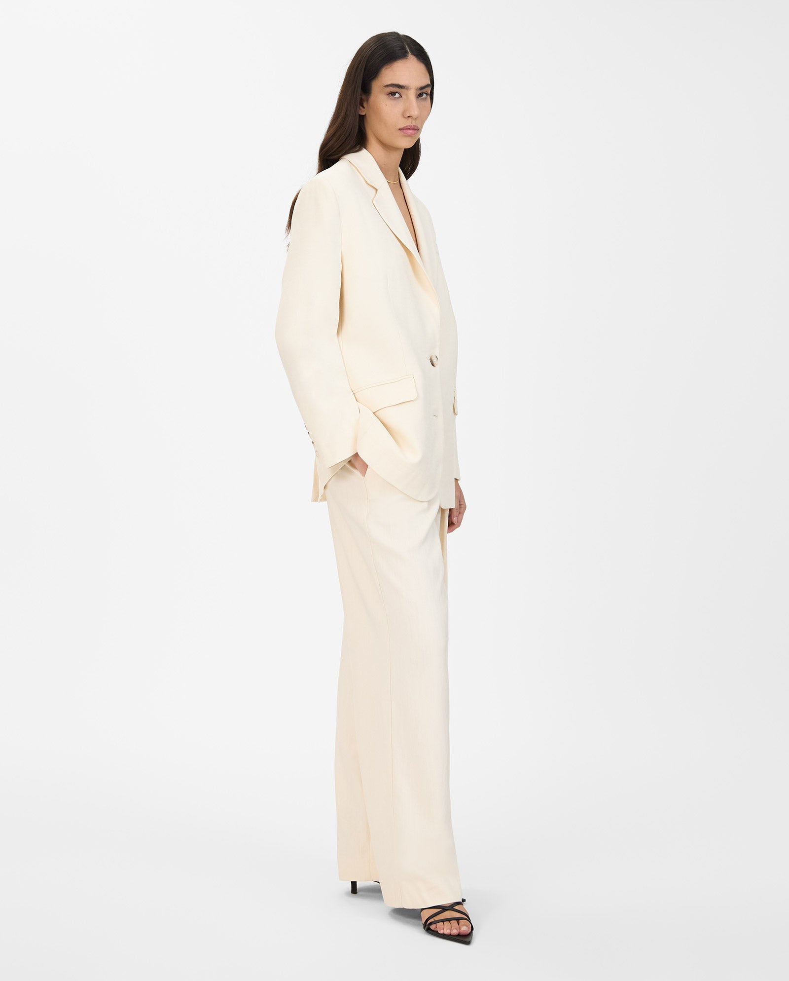 A woman stands against a white background wearing the IVY OAK JUNA Blazer—an oversized, single-breasted cream blazer—styled with wide-leg pants and black sandals.