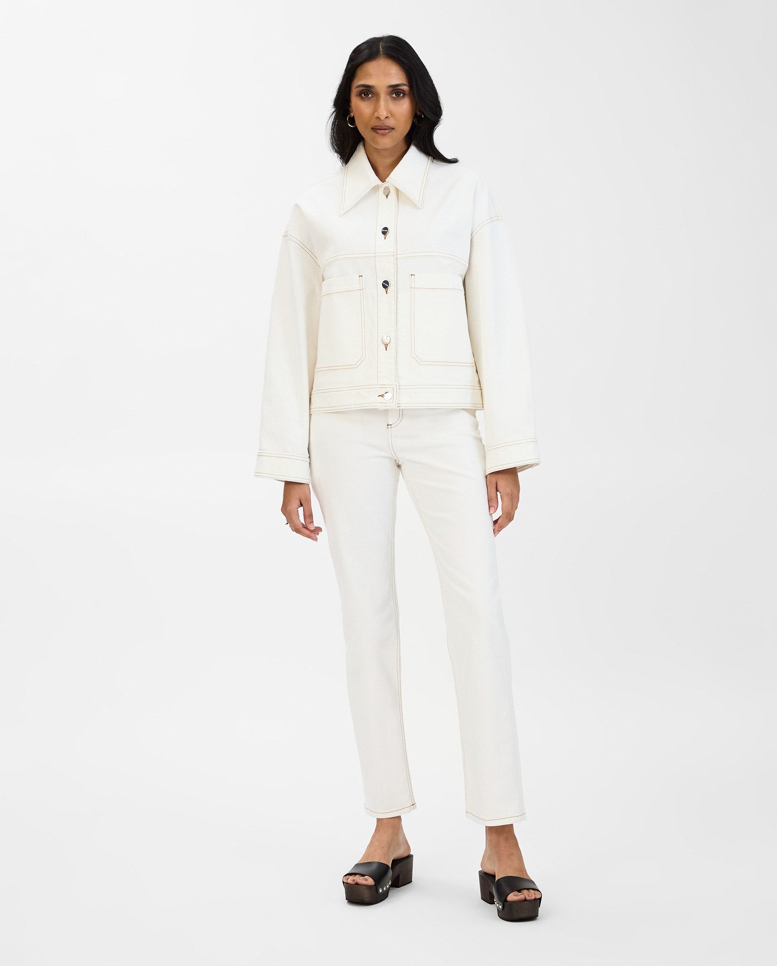 A woman stands on a white background wearing the IVY OAK JUPITER Jacket and matching white pants, paired with black platform sandals.