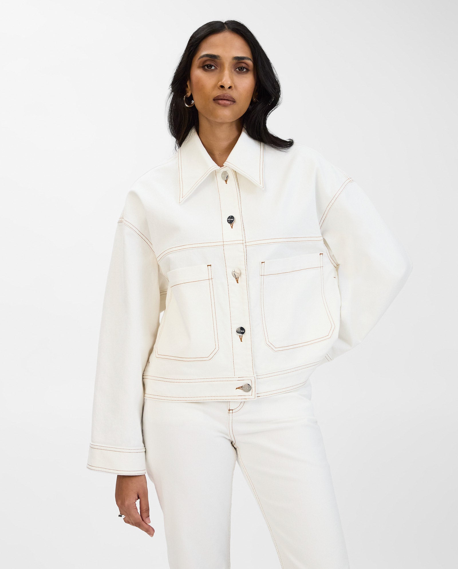 A woman poses with one hand on her hip, wearing the IVY OAK JUPITER cropped off-white denim jacket and matching pants against a plain background.