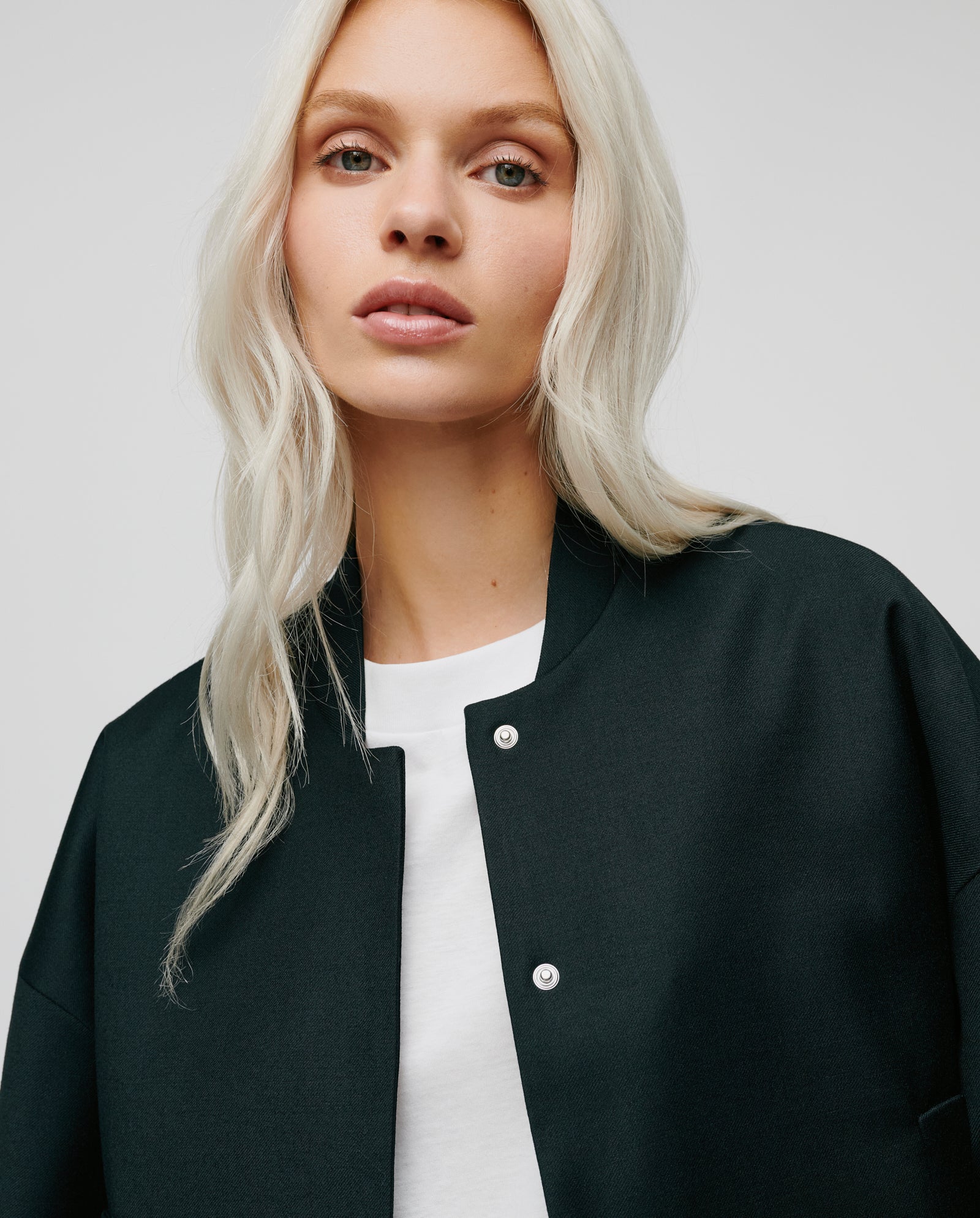 A woman with long blonde hair models the IVY OAK JAMIE Jacket—an oversized black bomber—over a white top, gazing at the camera for a relaxed vibe.