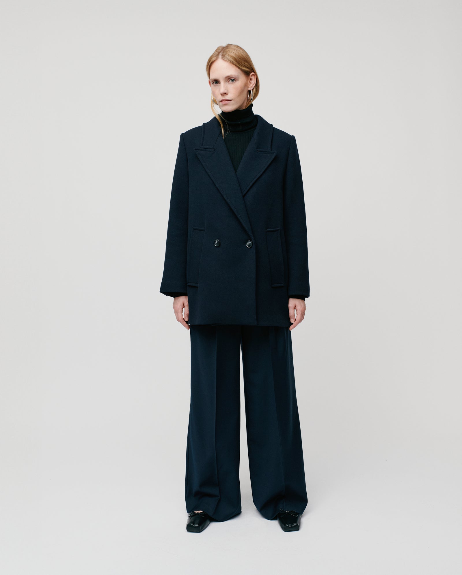 A person stands against a plain background wearing the IVY OAK JENNA double-breasted jacket, wide-leg pants, and a black turtleneck.
