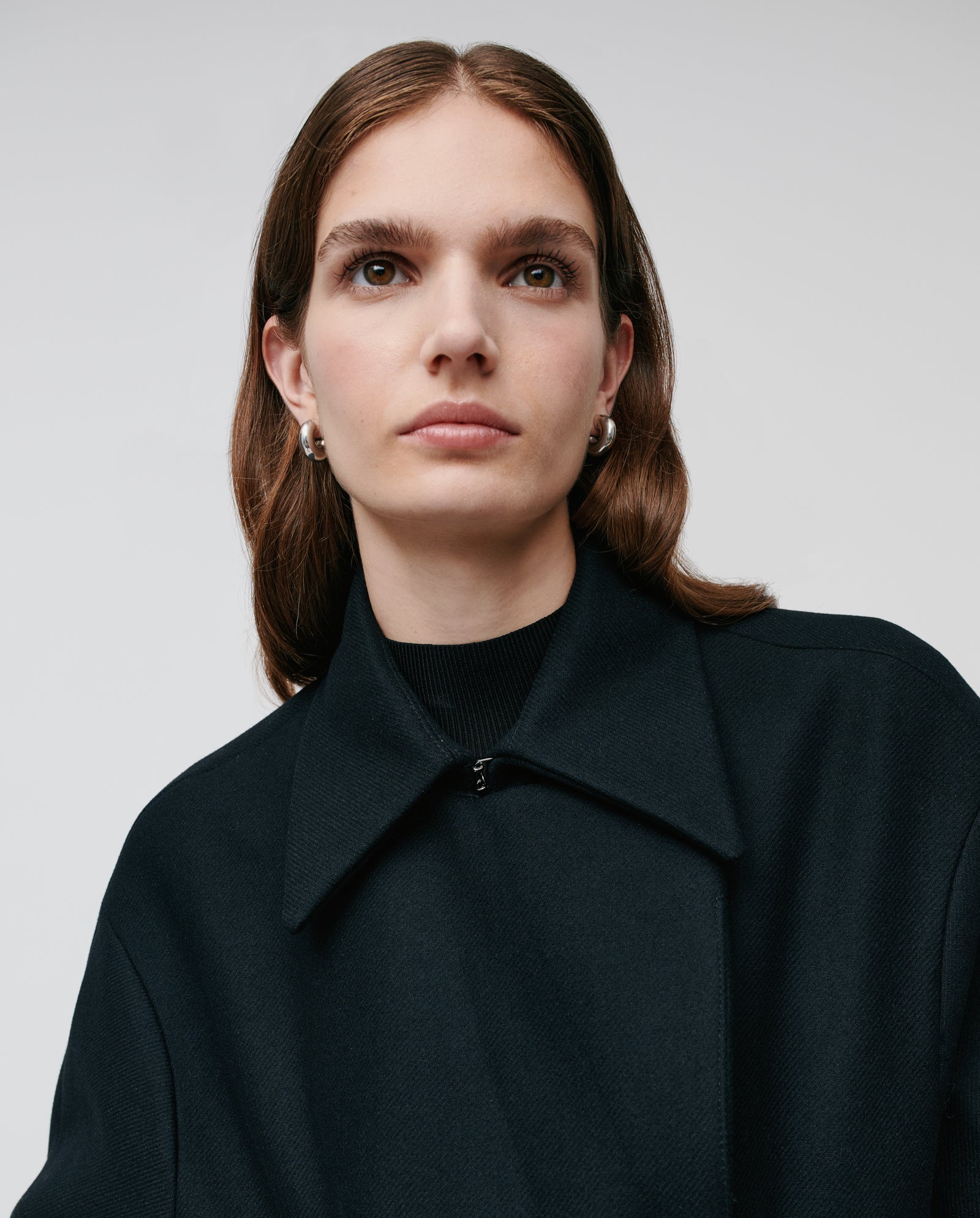 Woman with long brown hair wearing the black JANET Jacket by IVY OAK and pearl earrings, looking slightly to the side.