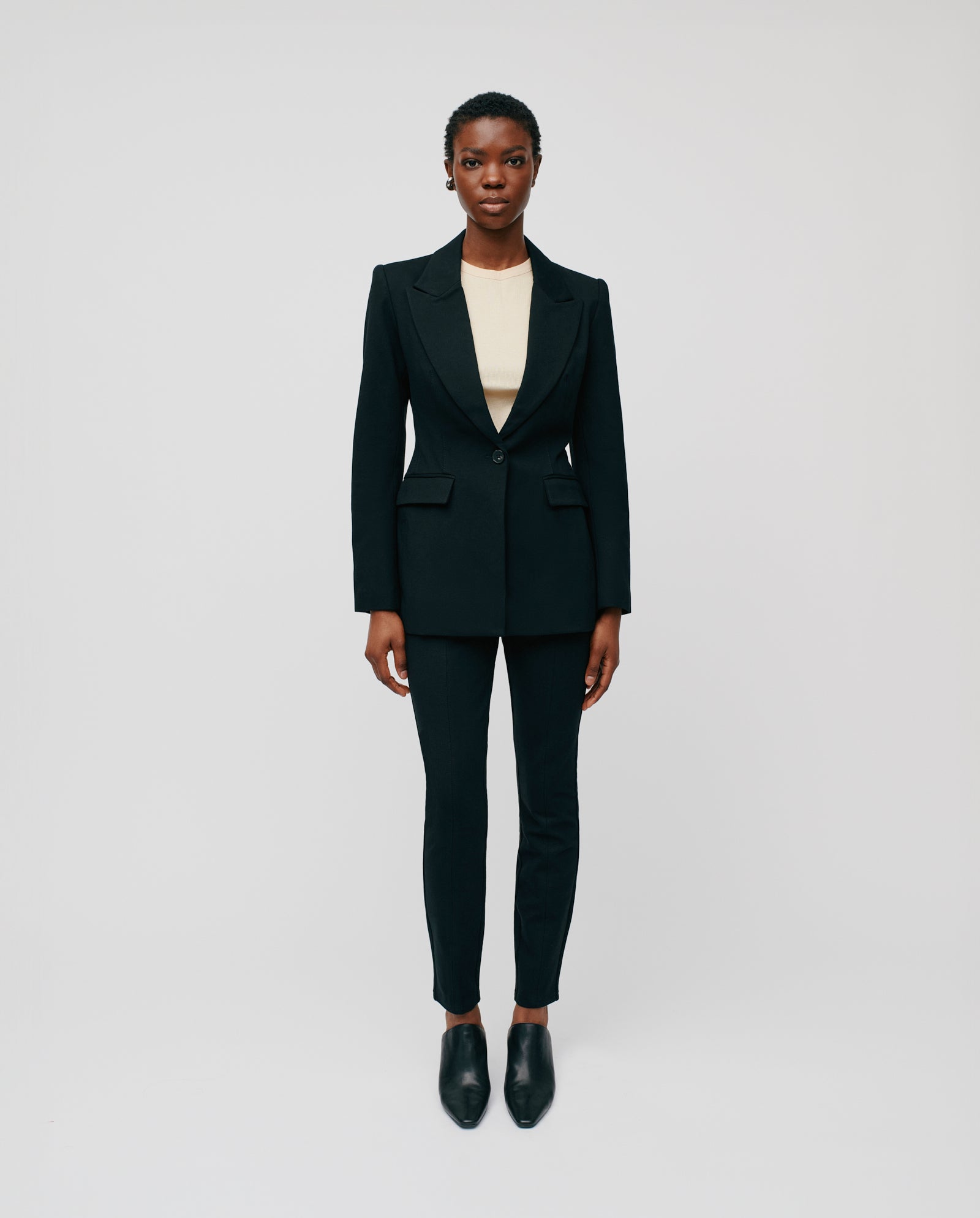 A person stands facing forward in an IVY OAK JOLANKA KIM Blazer, which features an elegant silhouette with subtle topstitching, paired with a cream top and black shoes against a plain light background.