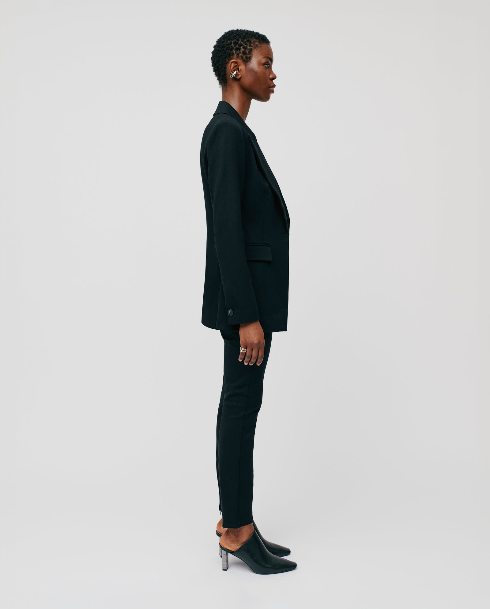A person in a black IVY OAK JOLANKA KIM Blazer stands in profile against a plain, light background, showcasing elegant topstitching and a refined silhouette with heeled shoes.