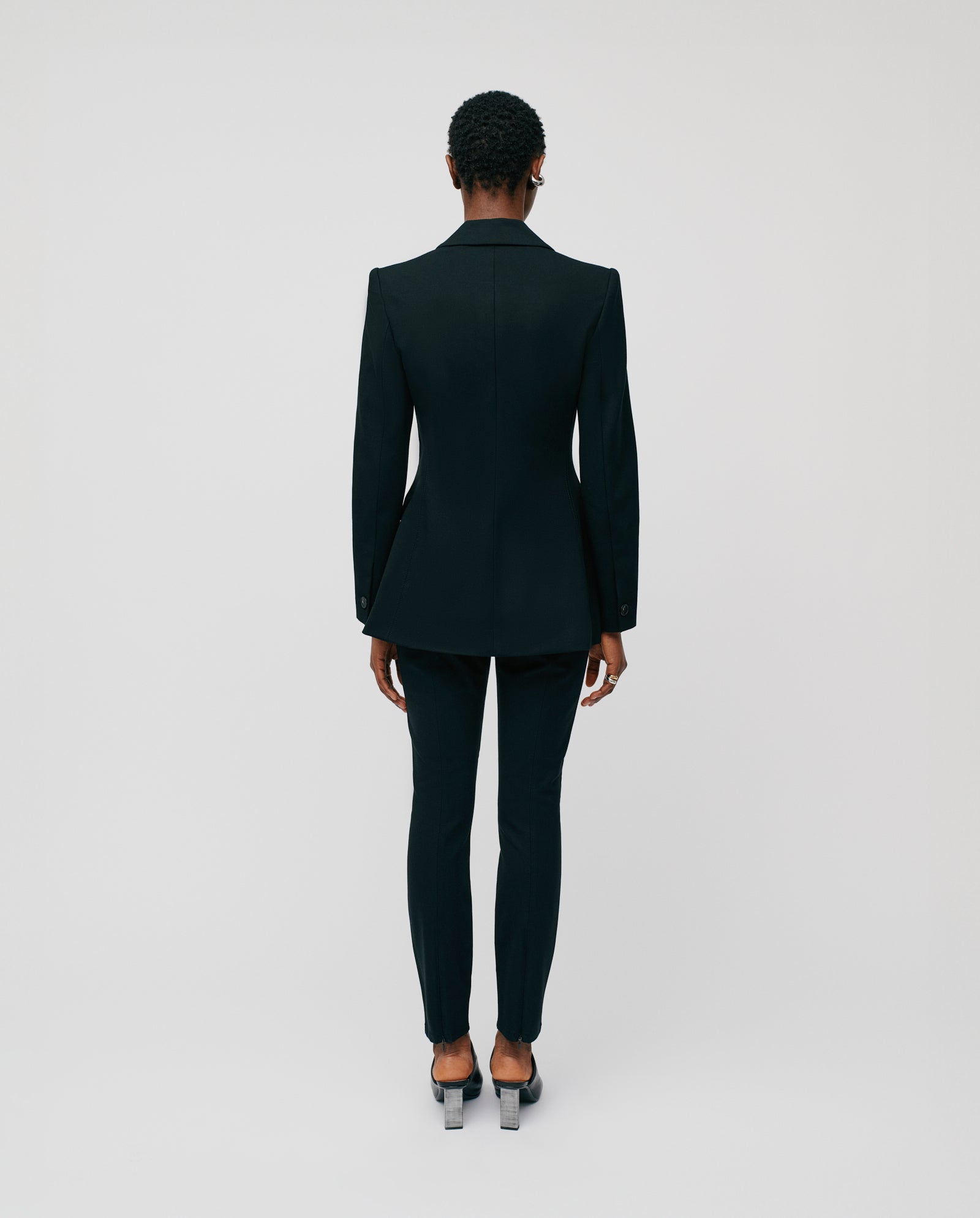 A person in a tailored black IVY OAK JOLANKA KIM Blazer and heels stands facing away against a plain light background, highlighting the elegant silhouette and precise topstitching of the blazer.