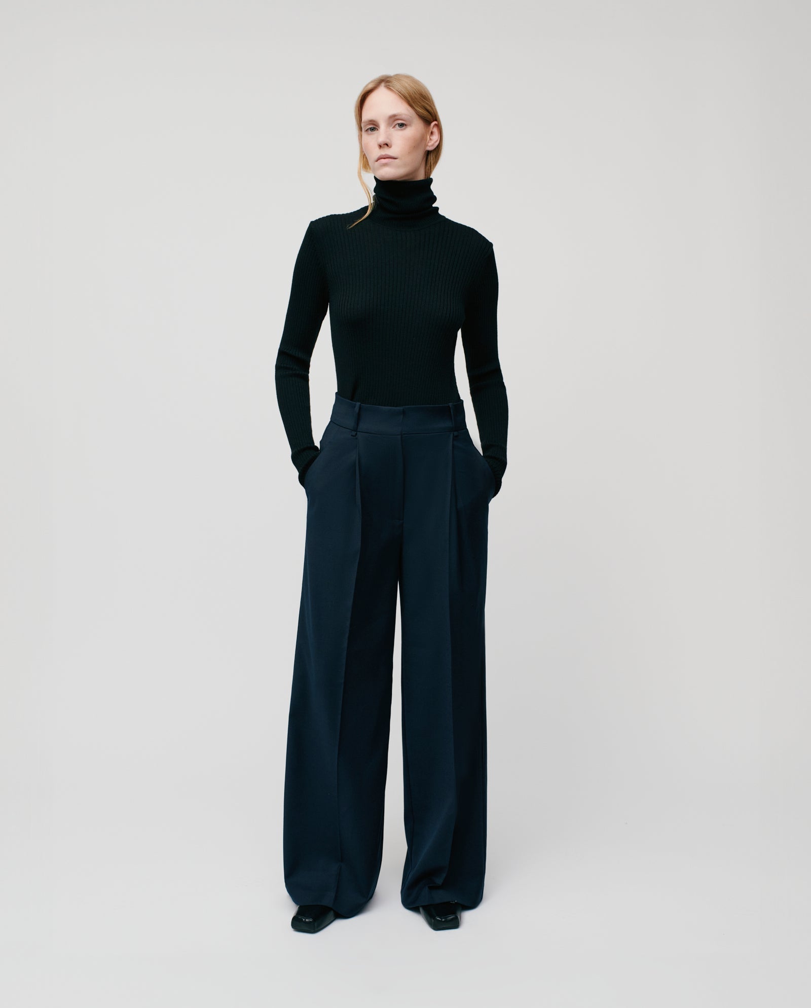 A woman in a black turtleneck and IVY OAK’s PRESCILLIA Trousers stands with hands in pockets, styled effortlessly for any occasion. Pair with the JOLANKA blazer for a refined look against a plain background.