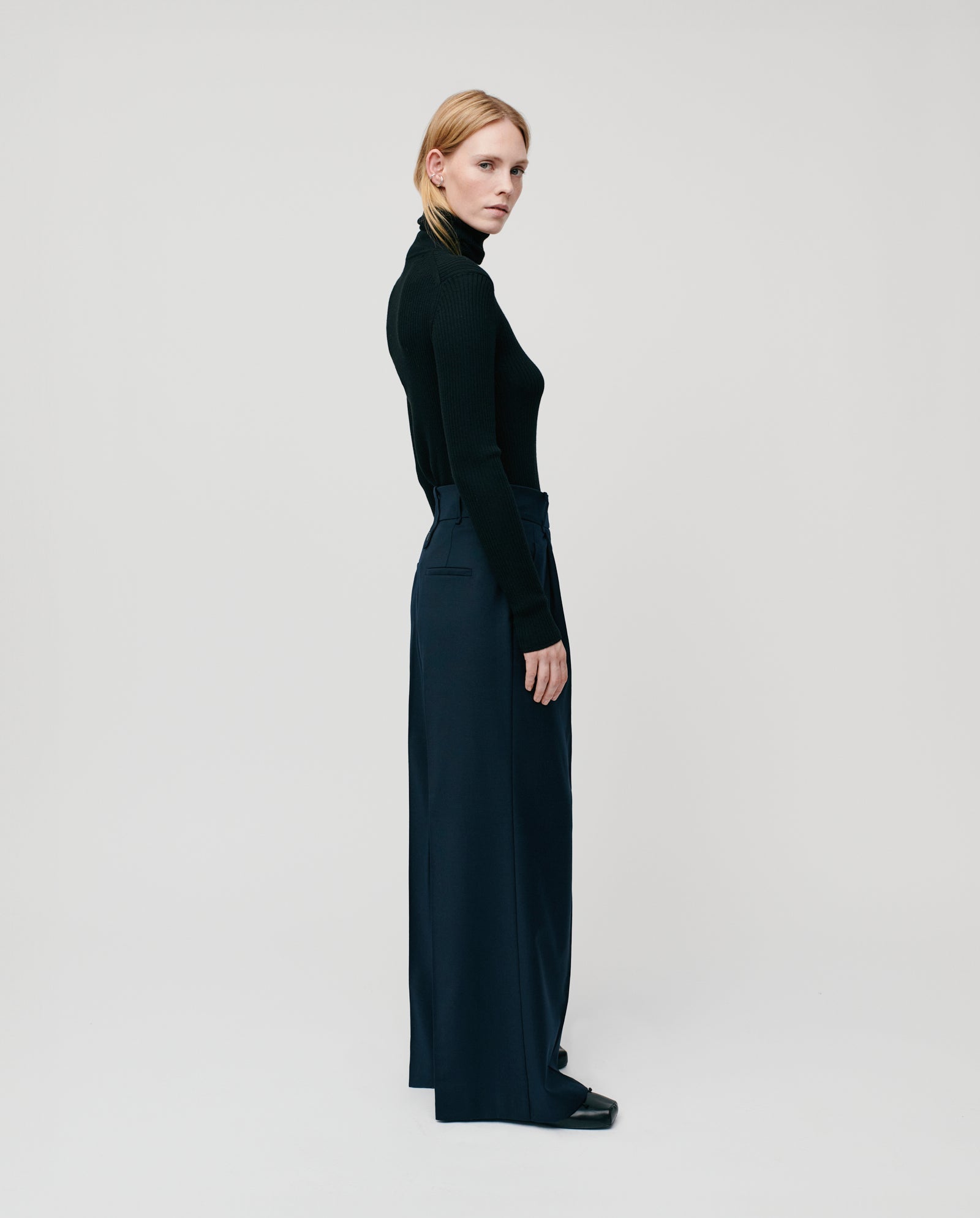 A woman in a black turtleneck and IVY OAK’s PRESCILLIA Trousers with adjustable length stands sideways against a plain white background.