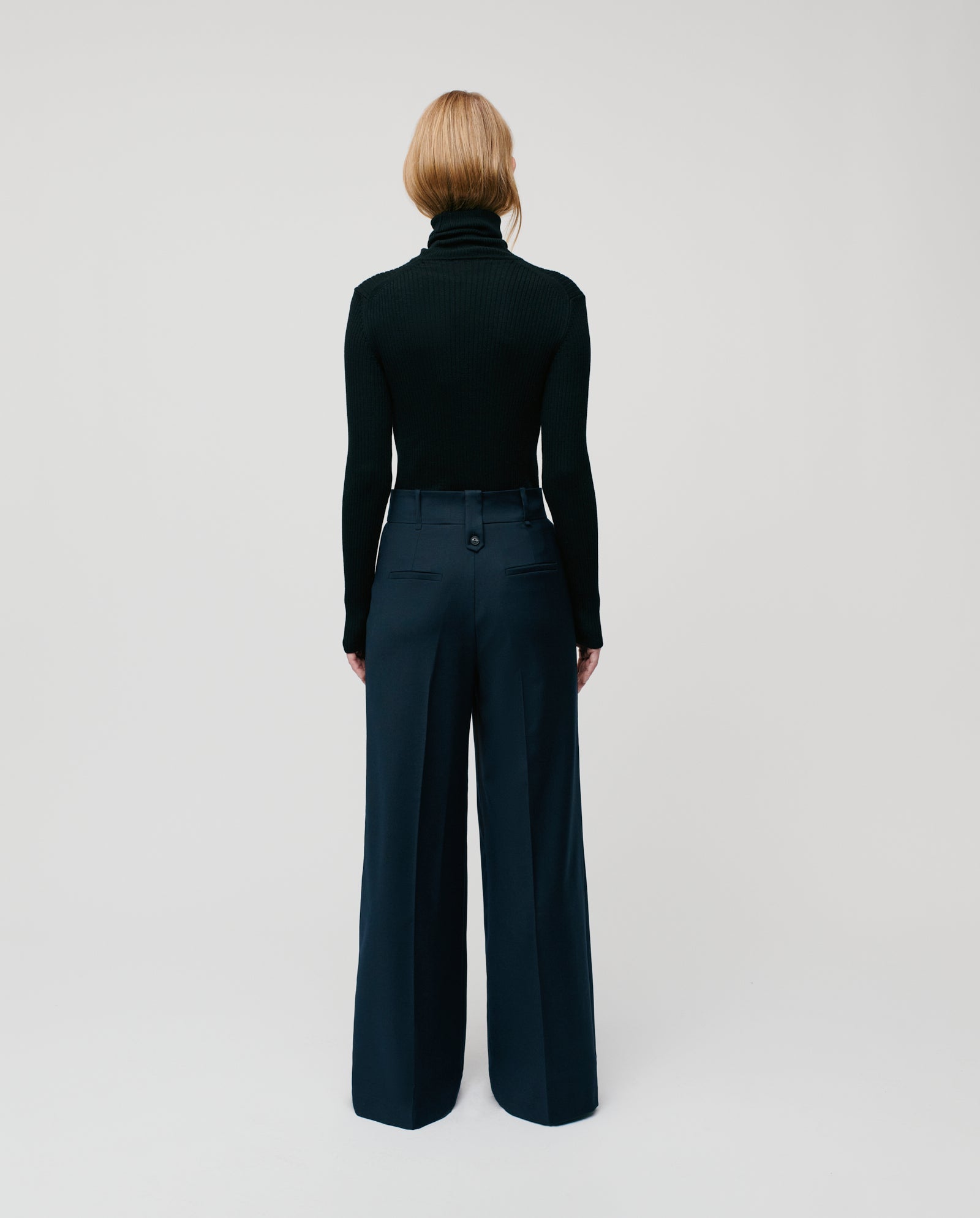 A person stands facing away against a plain background, wearing a black turtleneck, IVY OAK’s PRESCILLIA adjustable-length wide leg trousers, and a JOLANKA blazer.