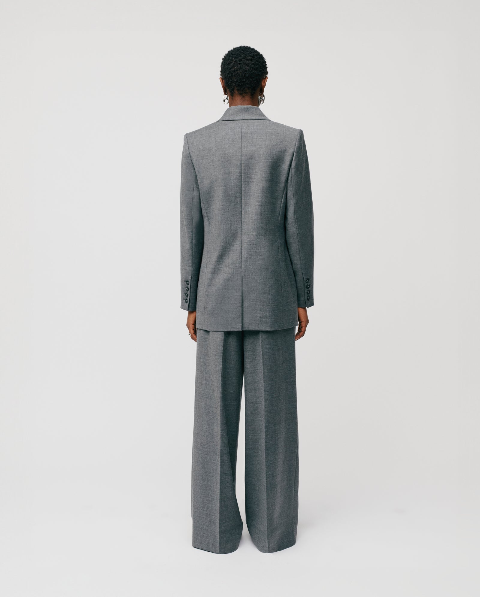 A person with short hair stands facing away against a plain white background, wearing the IVY OAK JULIETTE ROSE Blazer in gray with a boxy fit and button closure.