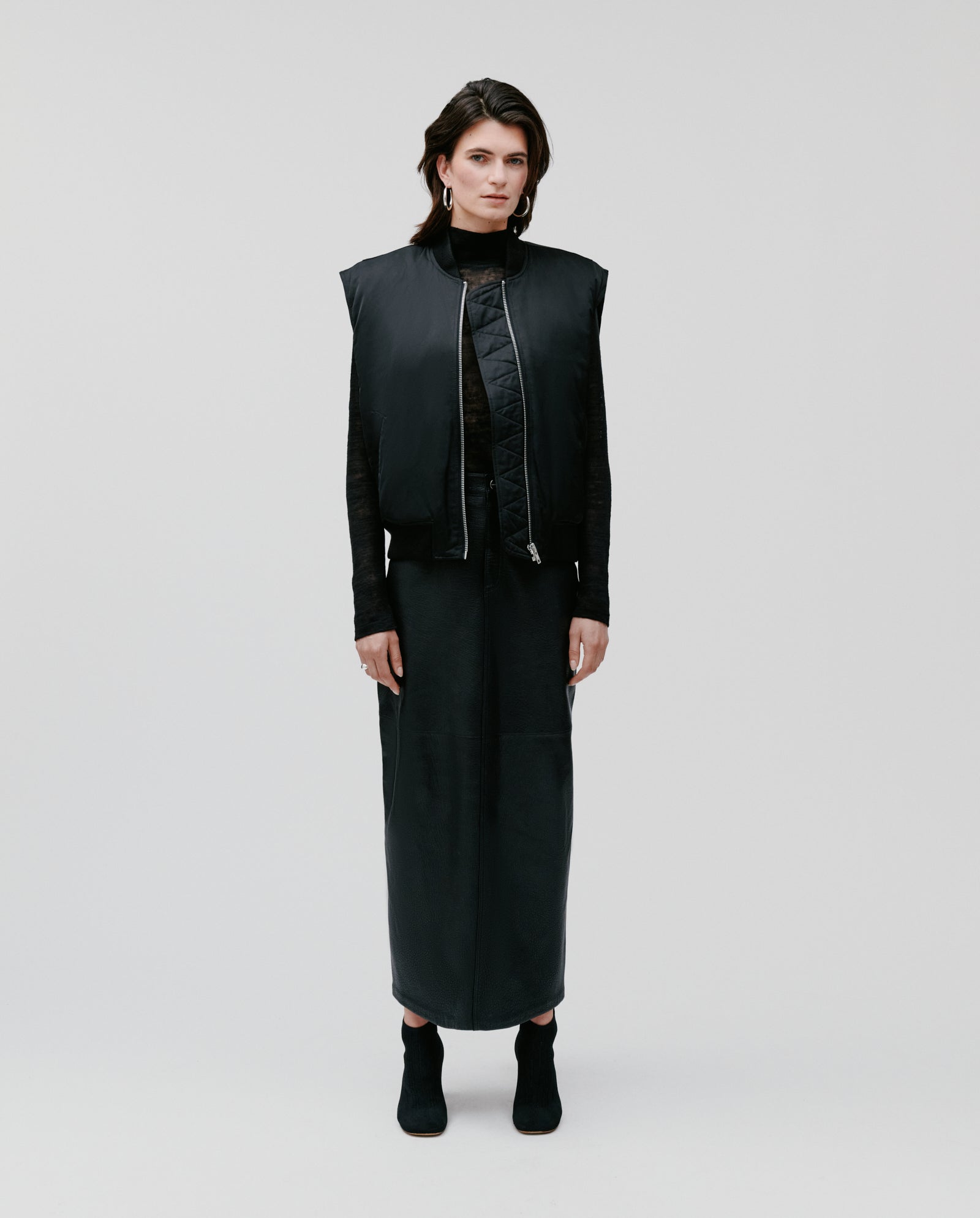 A woman wears the IVY OAK JAMILA Jacket, a loose-fitted black vest, paired with a long black skirt and black boots, standing against a plain light background.