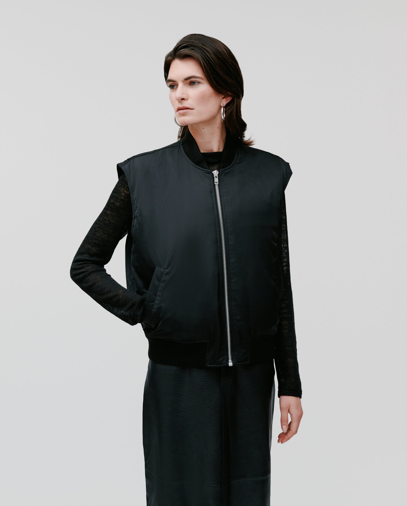 A person in the IVY OAK JAMILA Jacket, a loose-fitted black zip-up vest styled with a skirt and hand in pocket, stands against a plain background—ideal transitional outerwear for every season.