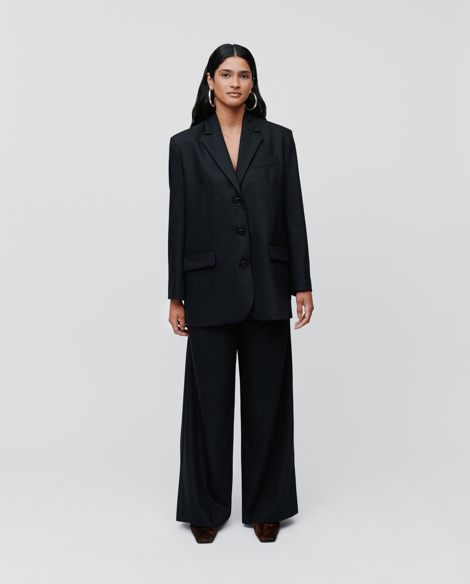 A woman stands against a plain light background wearing the IVY OAK JACQUELINE Blazer, a black oversized single breasted blazer paired with wide-leg pants for a boxy, modern look.