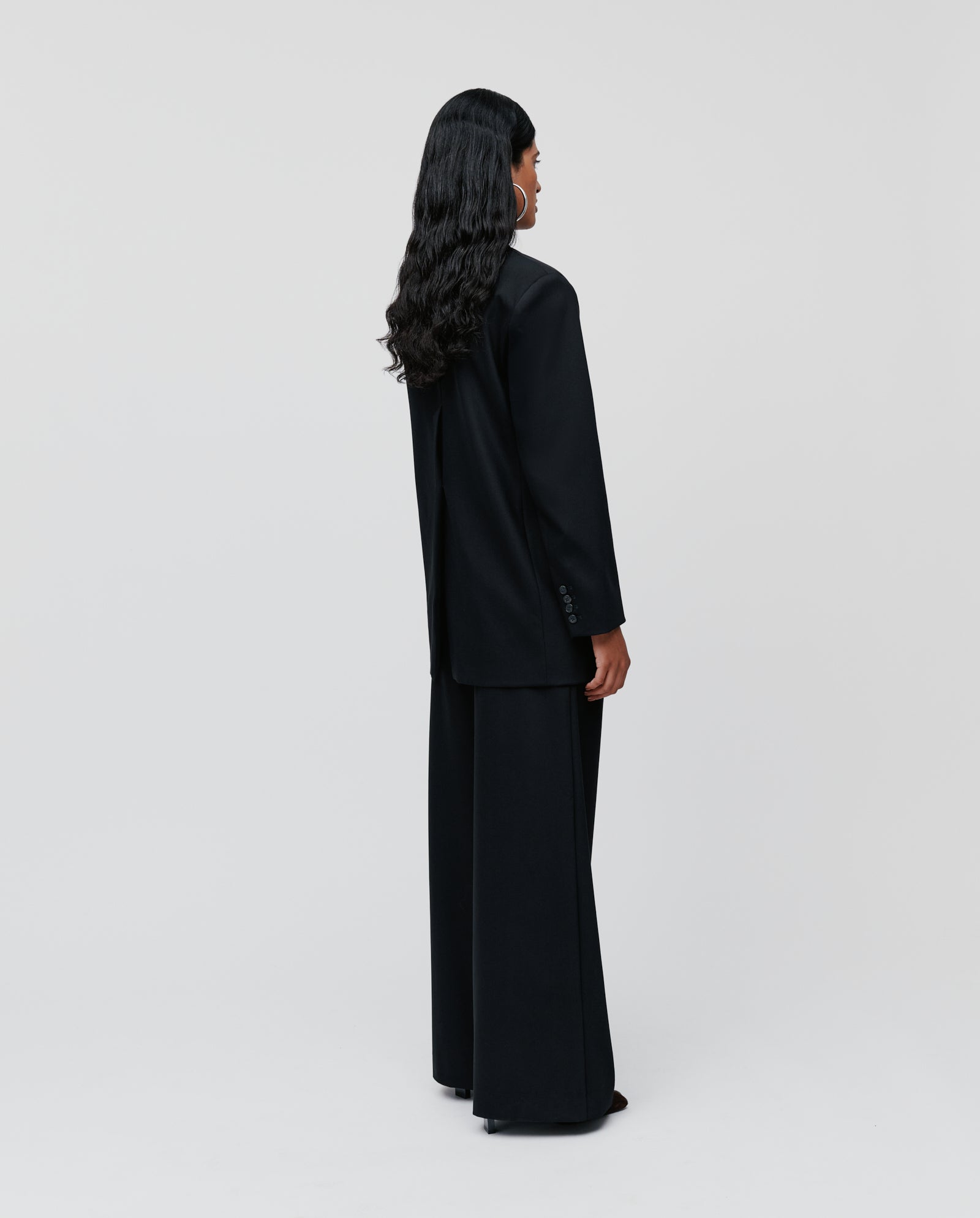 A person with long wavy hair, wearing the IVY OAK JACQUELINE Blazer in a black boxy fit, stands facing away against a plain white background.