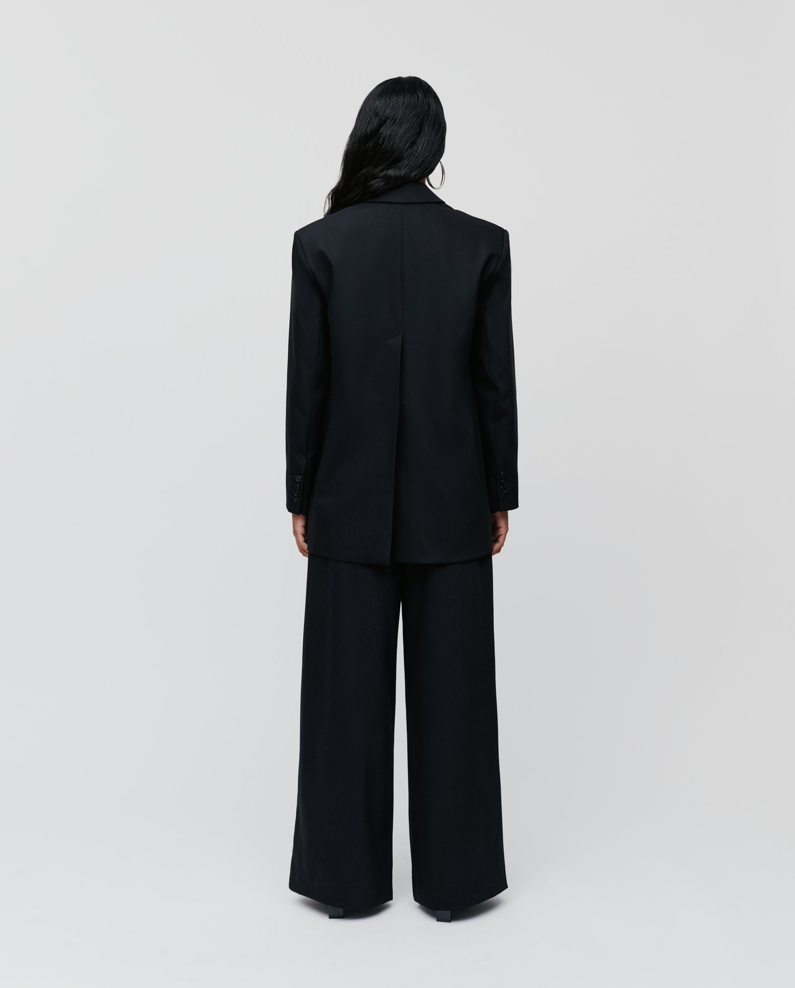 A person with long dark hair, wearing the IVY OAK JACQUELINE Blazer—a black, boxy single-breasted blazer—stands facing away against a plain white background.