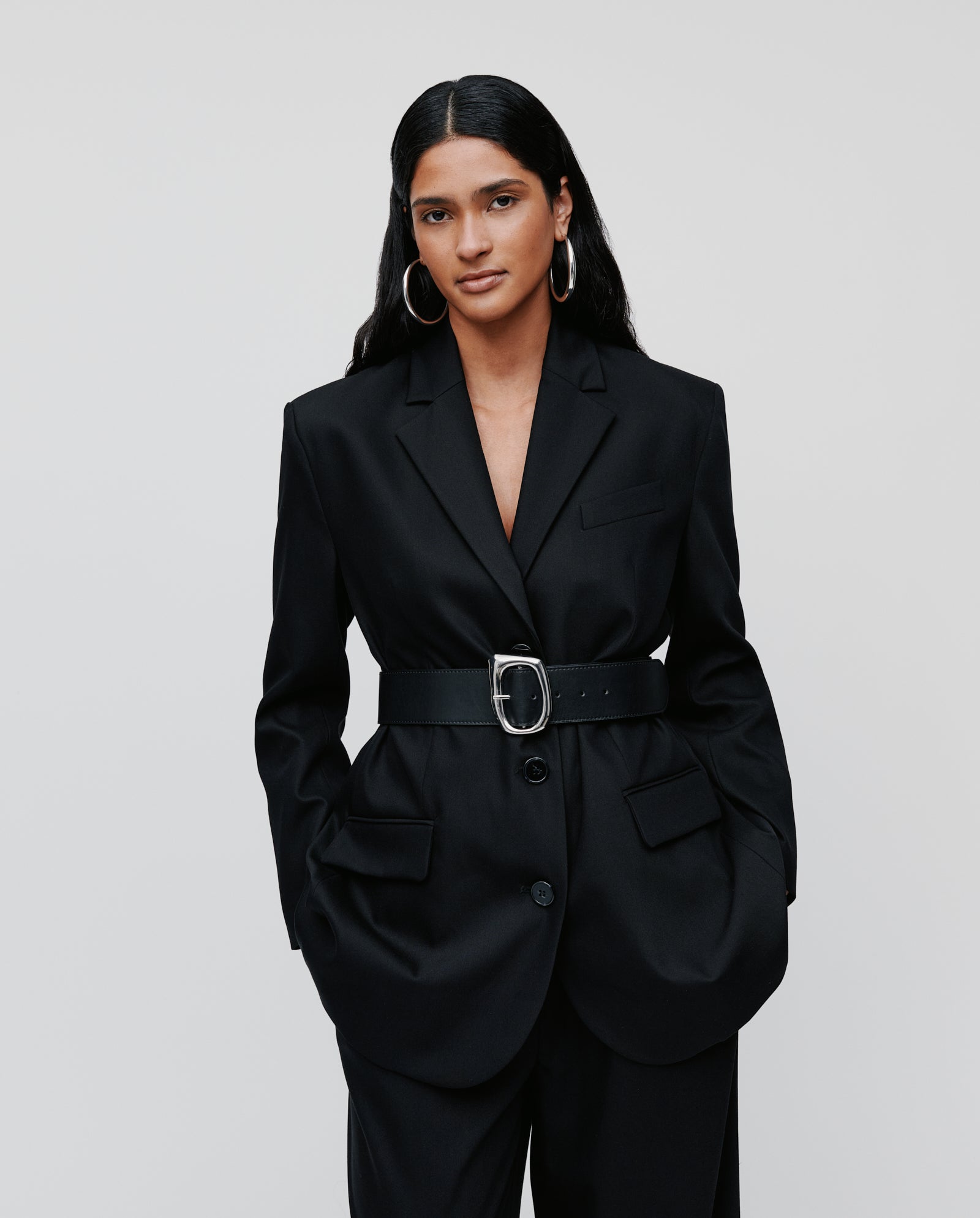 A woman wears the IVY OAK JACQUELINE Blazer—black, single-breasted, boxy fit with a wide belt—hands in pockets, standing against a plain background.