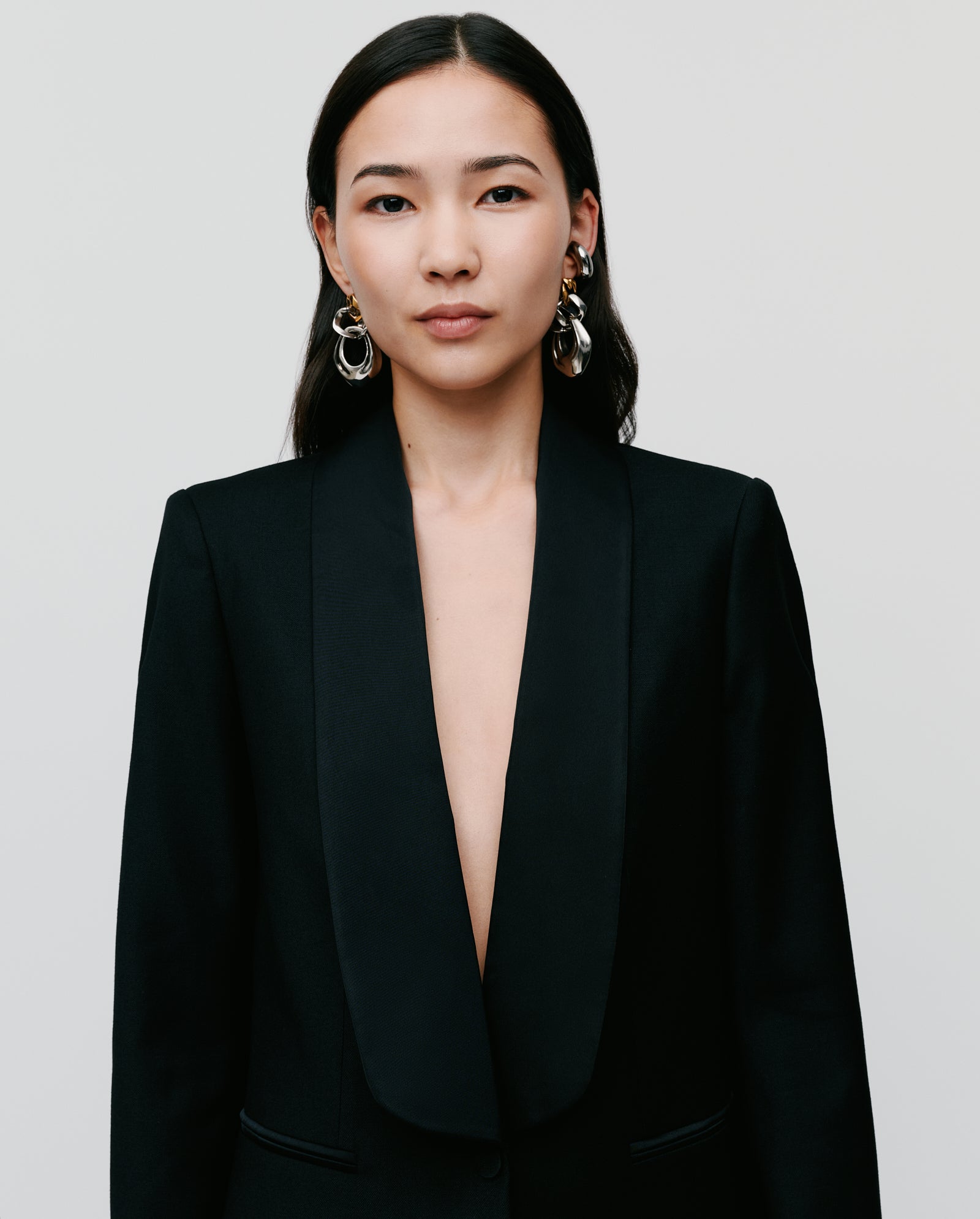 A woman with long dark hair wears the IVY OAK JANA MIA Blazer, a regular fit black shawl collar blazer with a front button closure, paired with statement earrings, posing against a plain background.