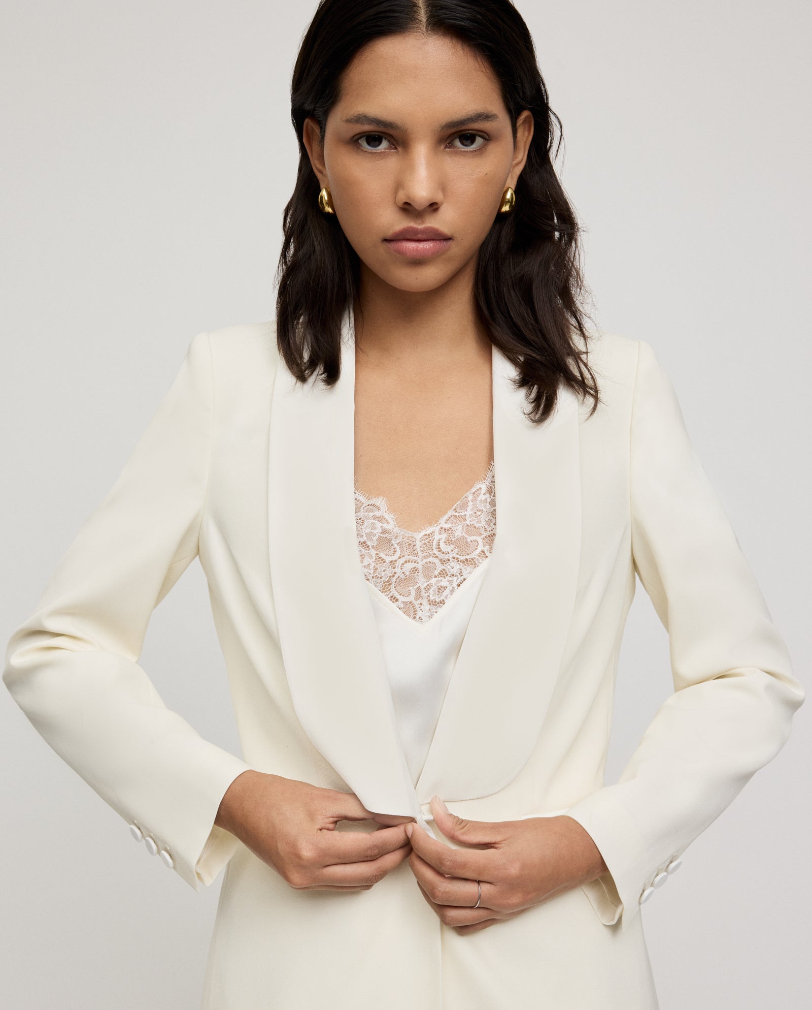 A woman wears the IVY OAK JANA MIA Blazer in a regular fit with a shawl collar and front button closure, pairing it with a lace top underneath, as she buttons up against a plain background.