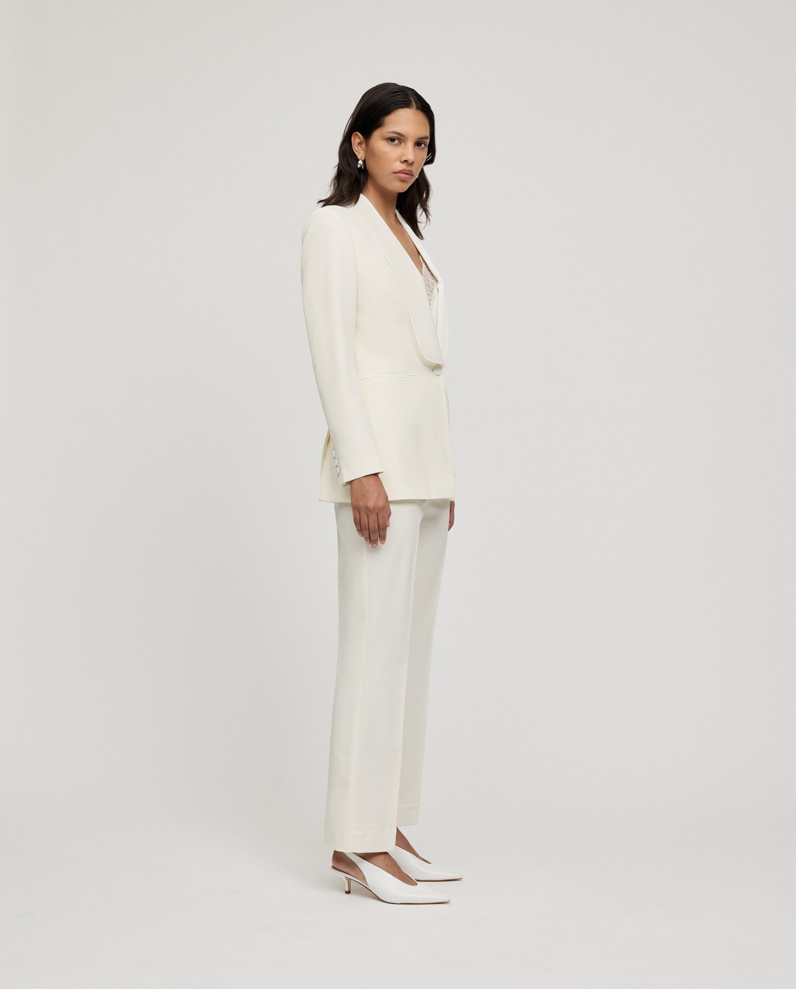 A woman in IVY OAK’s POLINA ROSE Trousers stands sideways against a plain light background, the sharp crease of her tailored pantsuit enhancing her polished look.