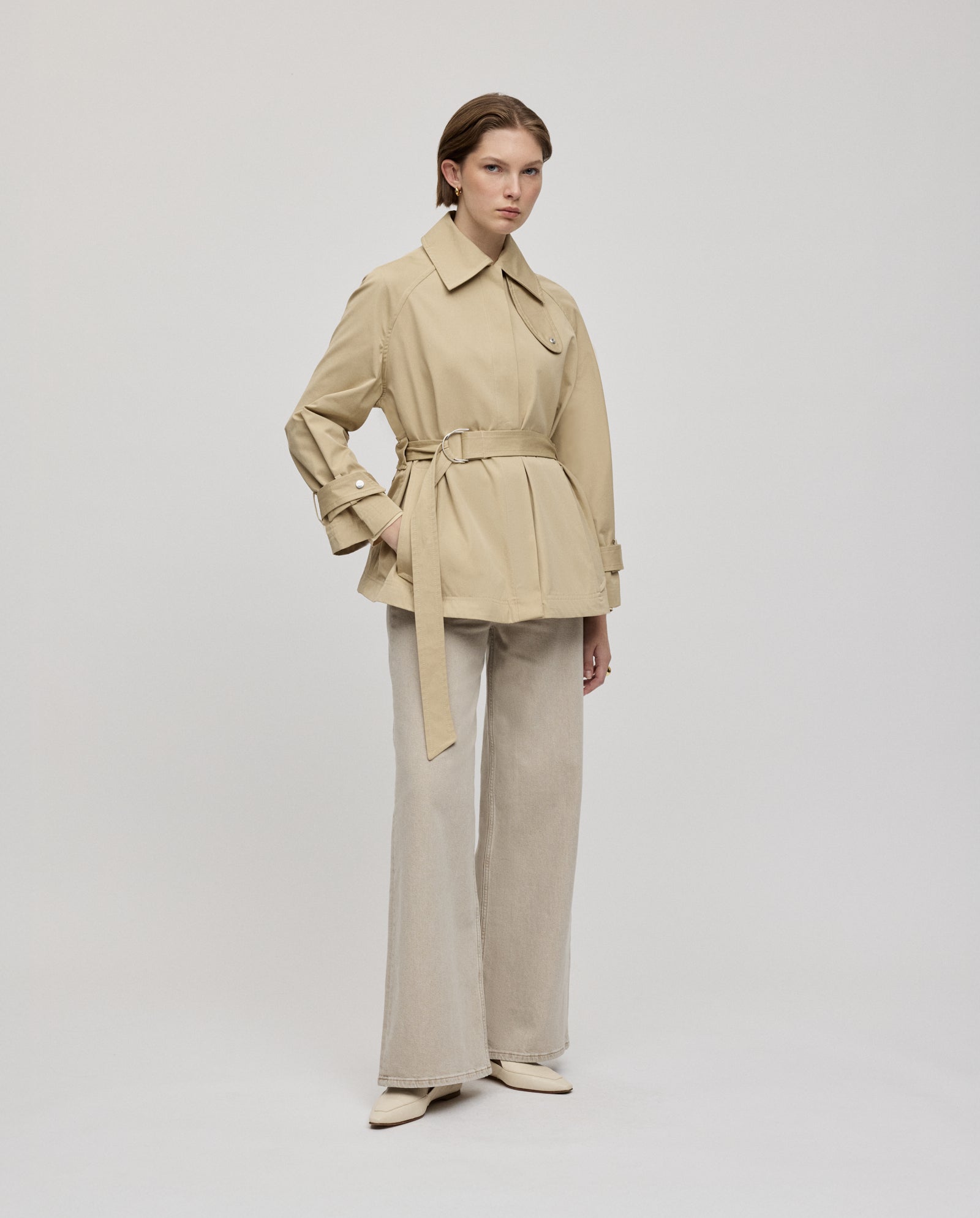 A woman stands against a plain background wearing the IVY OAK JULES Jacket in beige, styled with wide-leg light pants and cream shoes.
