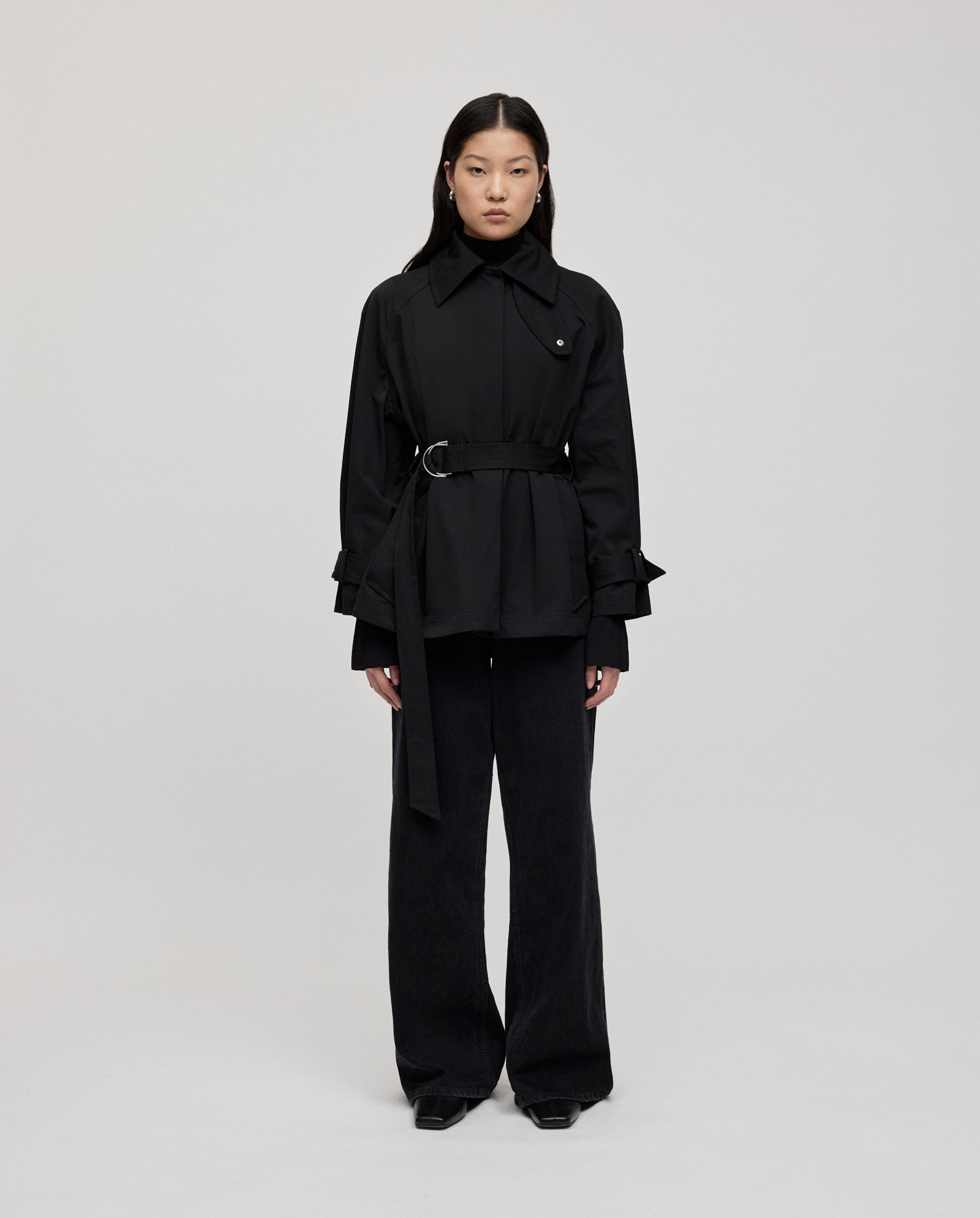 A woman faces forward wearing the IVY OAK JULES Jacket, a black trench with a belted waist, paired with black wide-leg pants against a plain backdrop.