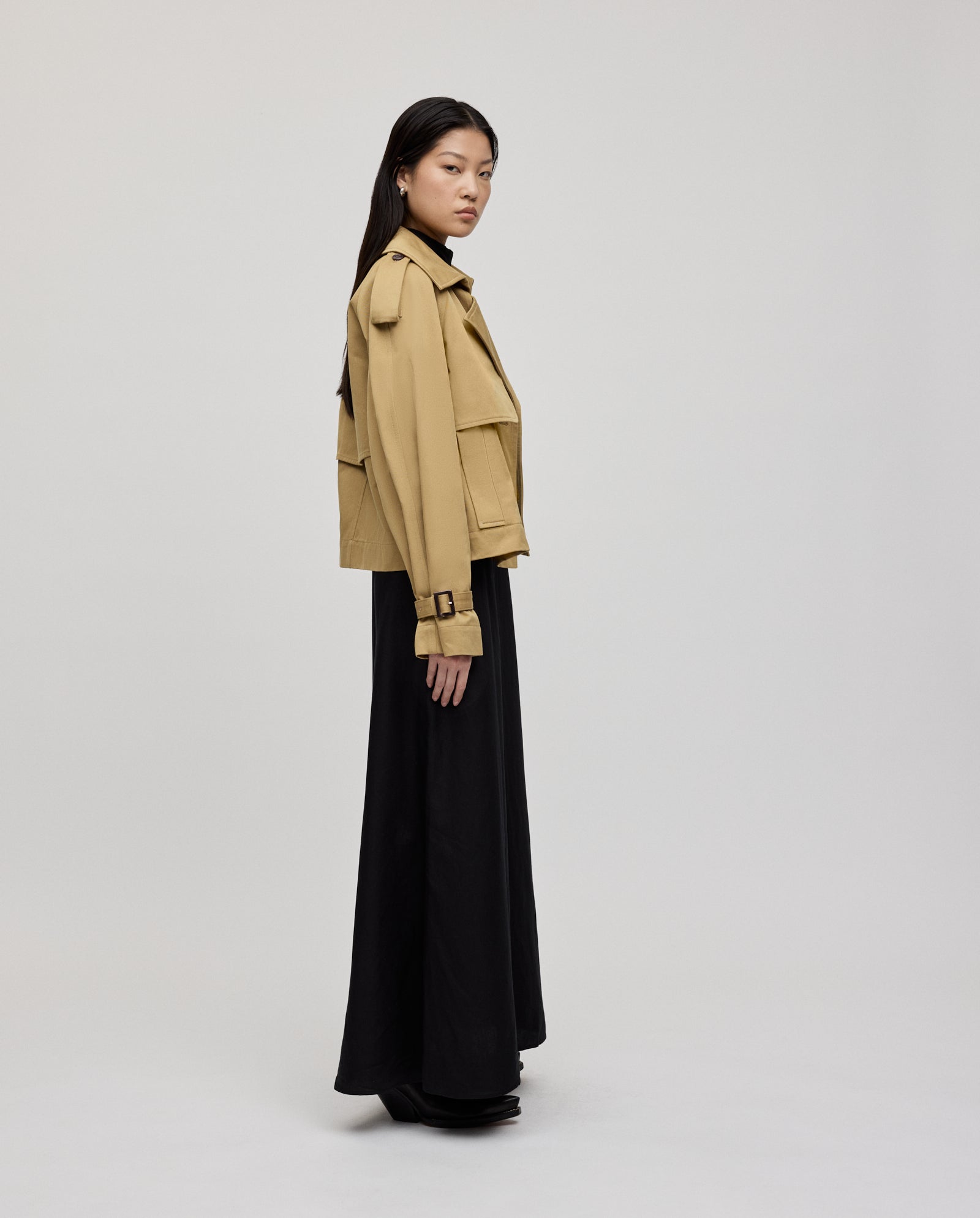 A woman wearing the IVY OAK JESSICA Jacket, a tan cropped trench coat with epaulettes and storm flaps, paired with a long black skirt, stands sideways against a plain light background.