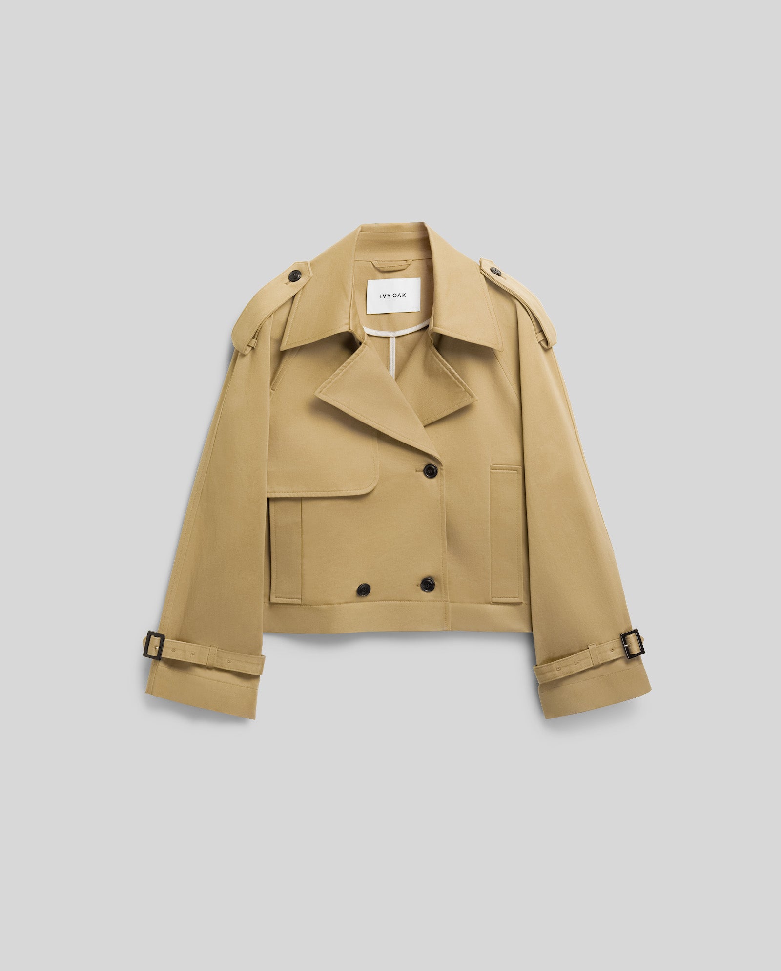 The JESSICA Jacket by IVY OAK is a beige cropped trench coat with wide lapels, front buttons, epaulettes, storm flaps, and buckled sleeve cuffs.