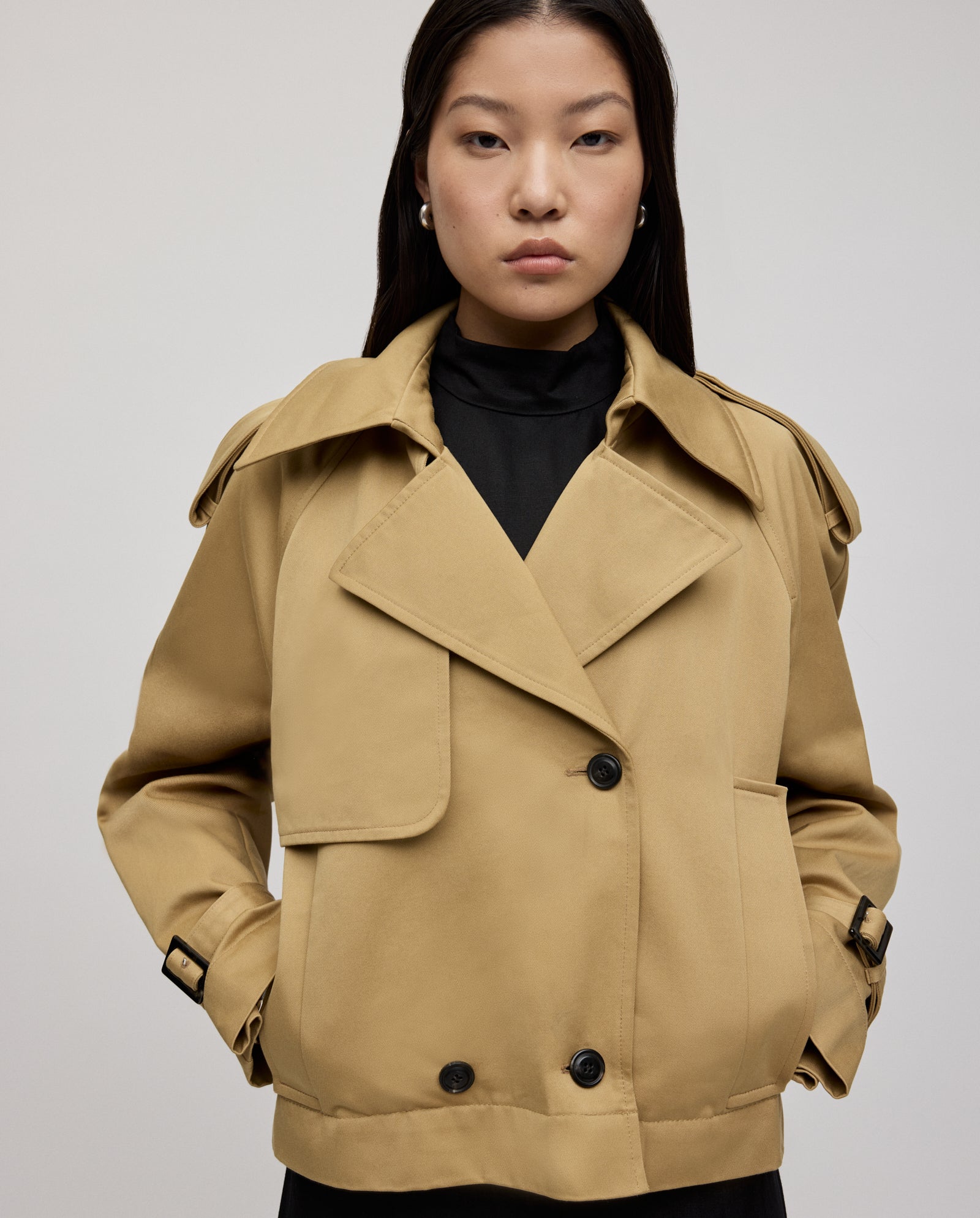 A woman wears the IVY OAK JESSICA Jacket, a tan double-breasted trench coat with black buttons and subtle epaulettes, standing against a plain background.