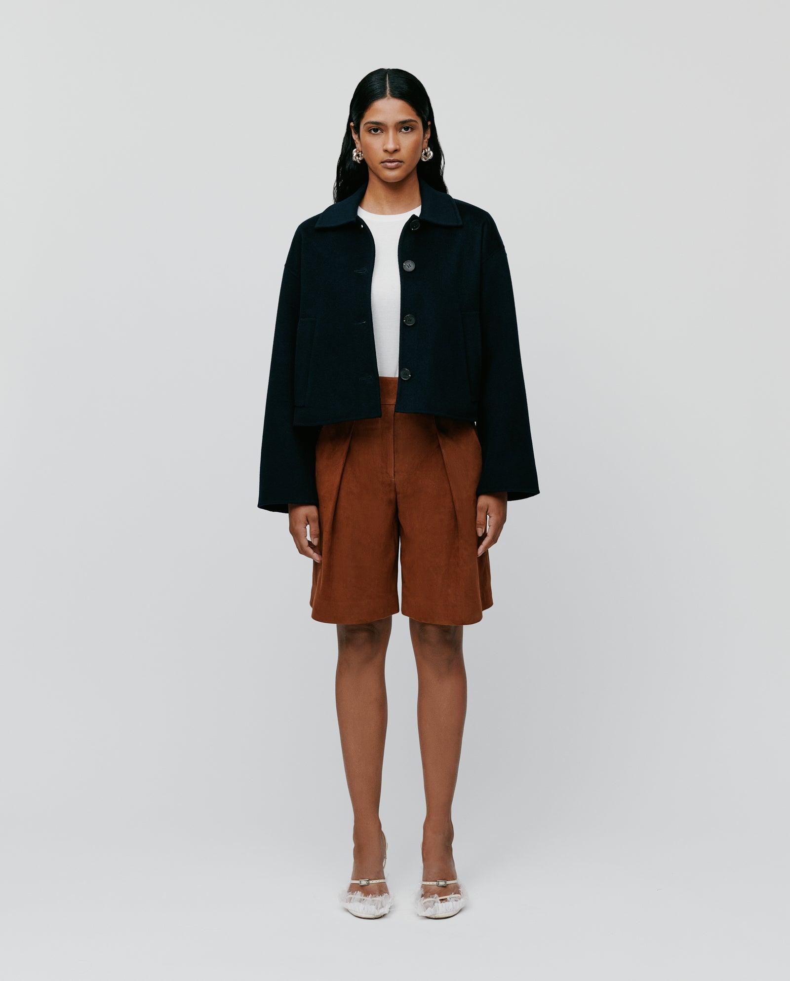 A woman poses against a plain background wearing the IVY OAK JAIA Wooljacket, paired with a white top, brown shorts, and transparent heels.