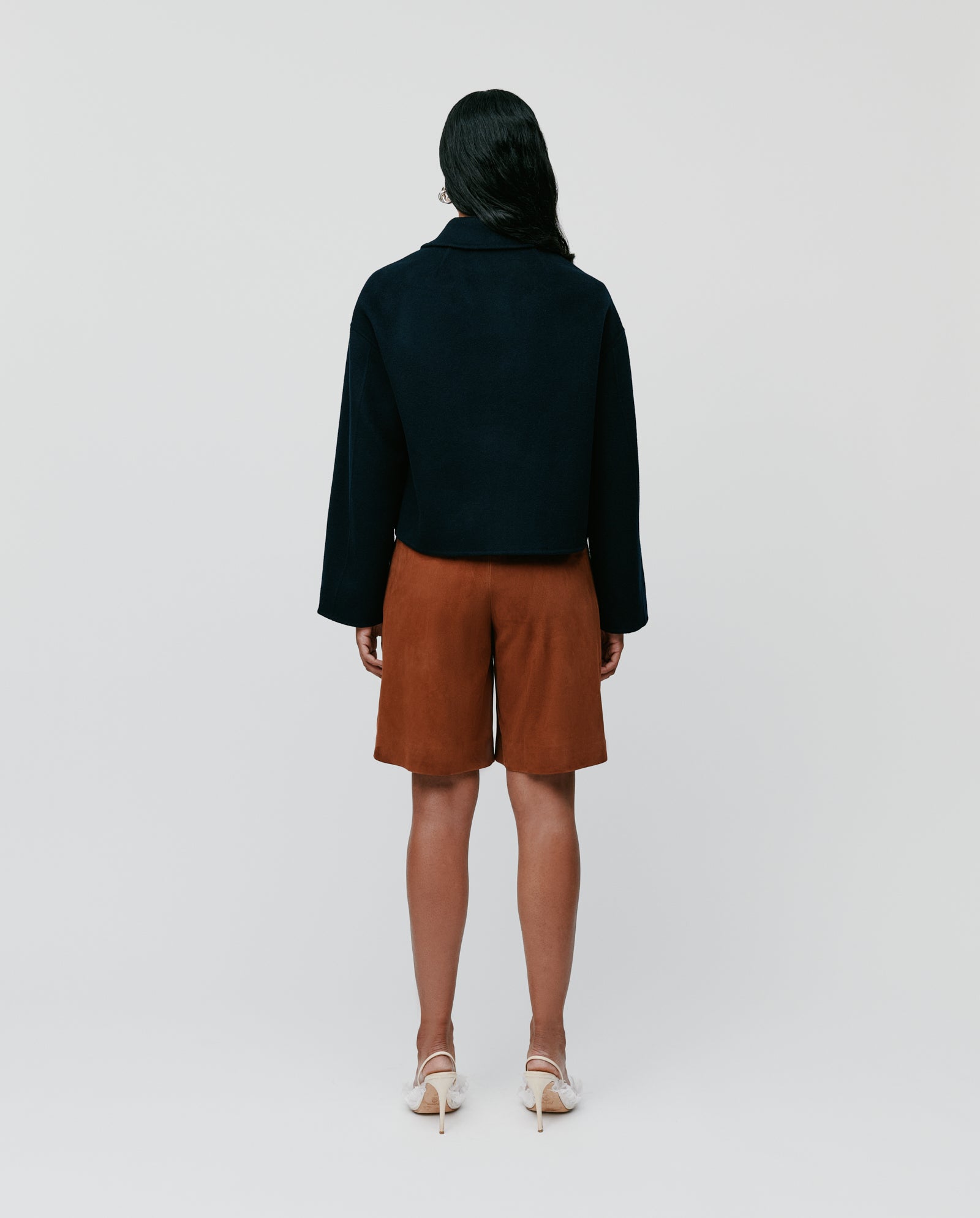 A person stands facing away, wearing the IVY OAK JAIA Wooljacket made from responsible wool, styled with brown shorts and white heels against a plain background.