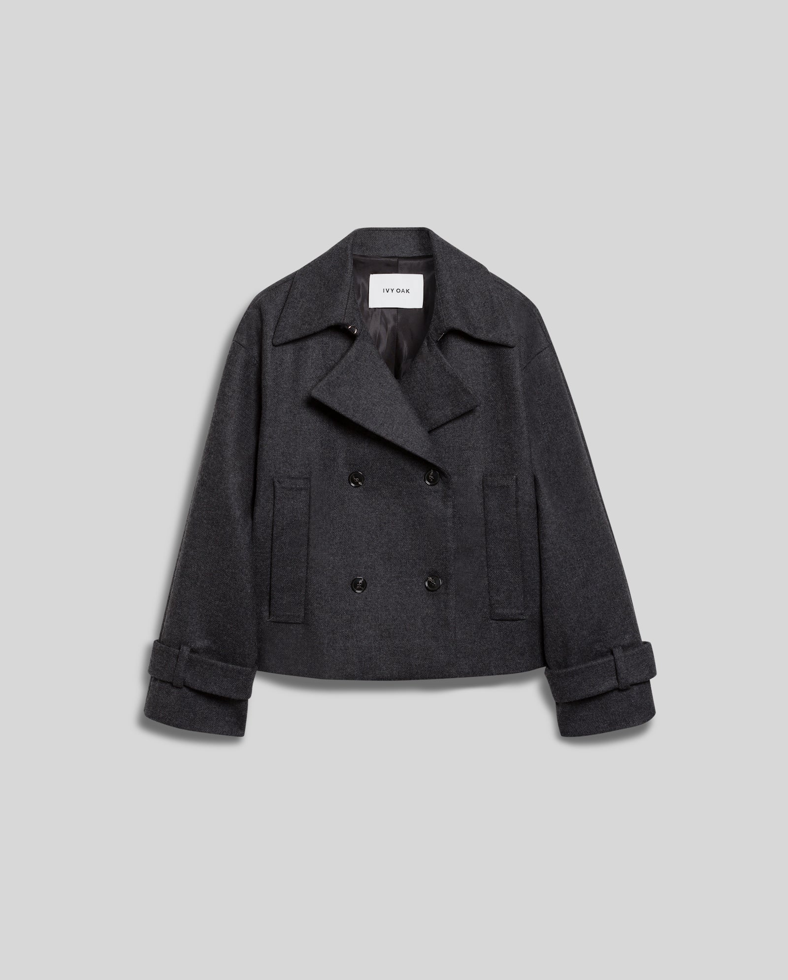 The JANET Wool Blazer by IVY OAK is a dark gray, double-breasted, cropped jacket in a cashmere-wool blend with wide lapels and cuffed sleeves, offering a modern trench look against a light gray background.