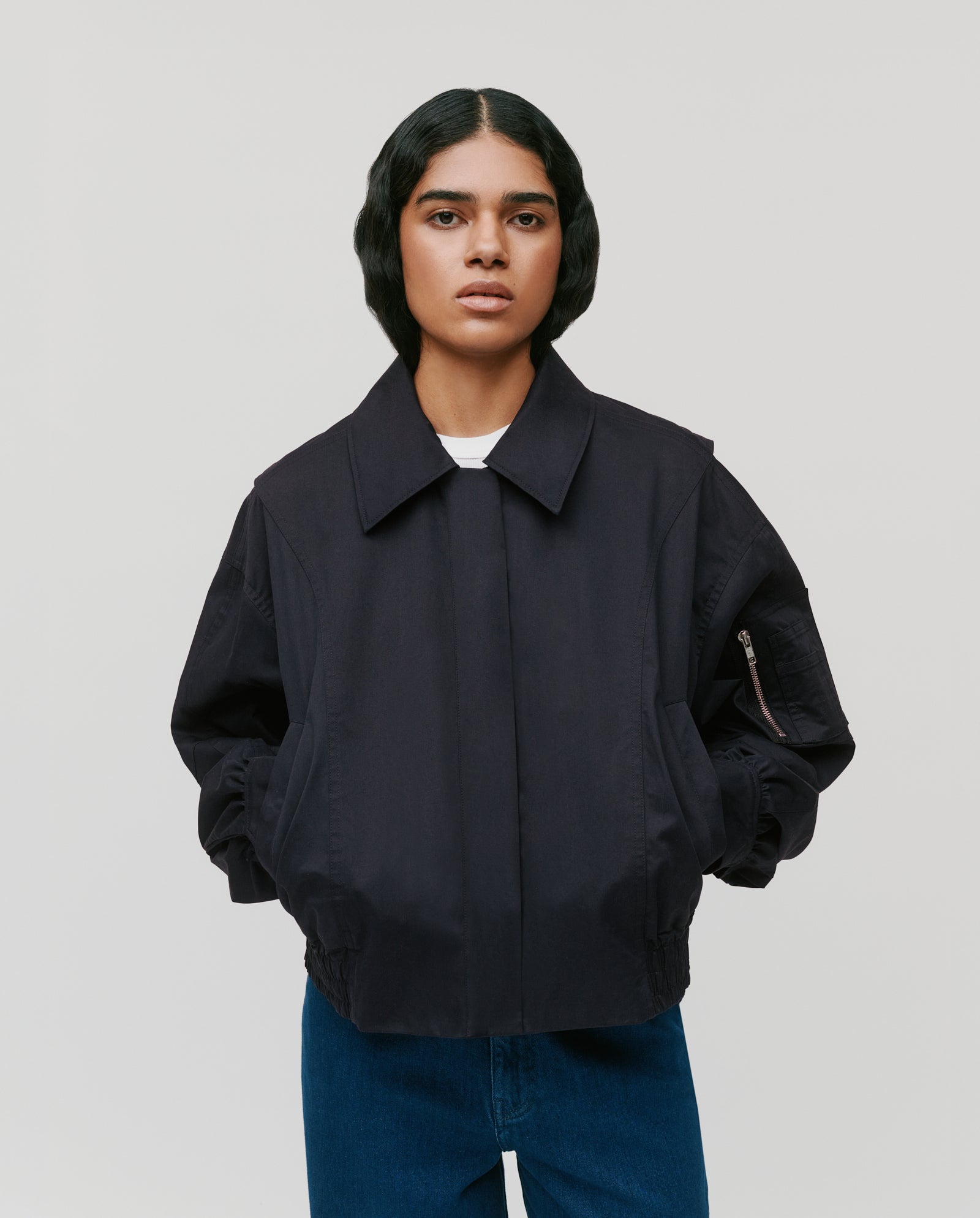 Model wearing the JANE ROSE Jacket by IVY OAK—a dark bomber style with a zipper—paired with blue jeans, standing hands in pockets against a plain background.