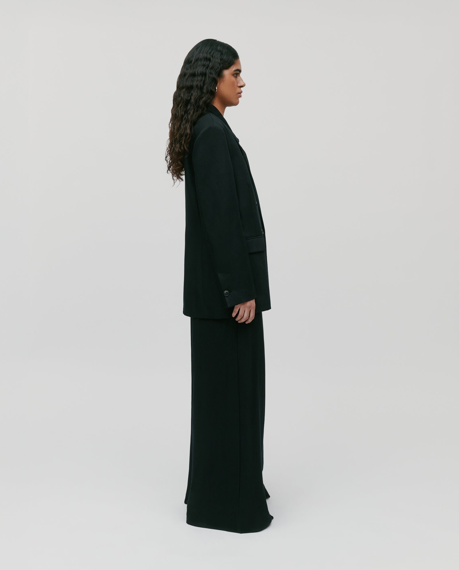 A person with long, dark hair stands in profile against a plain light background, wearing the JASEMINA ROSE Blazer by IVY OAK.