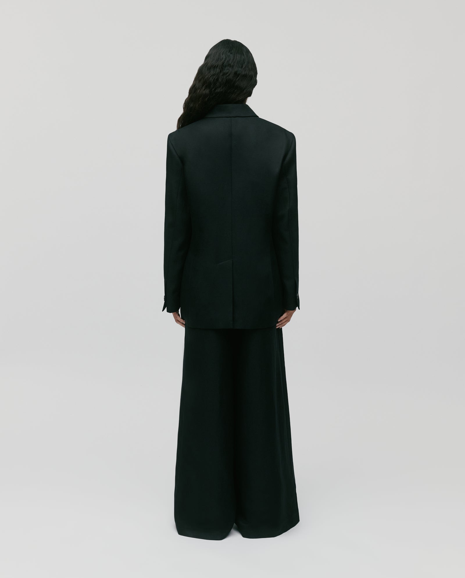 A person with long dark hair wears the IVY OAK JASEMINA ROSE Blazer in black linen, photographed from behind against a plain light background.