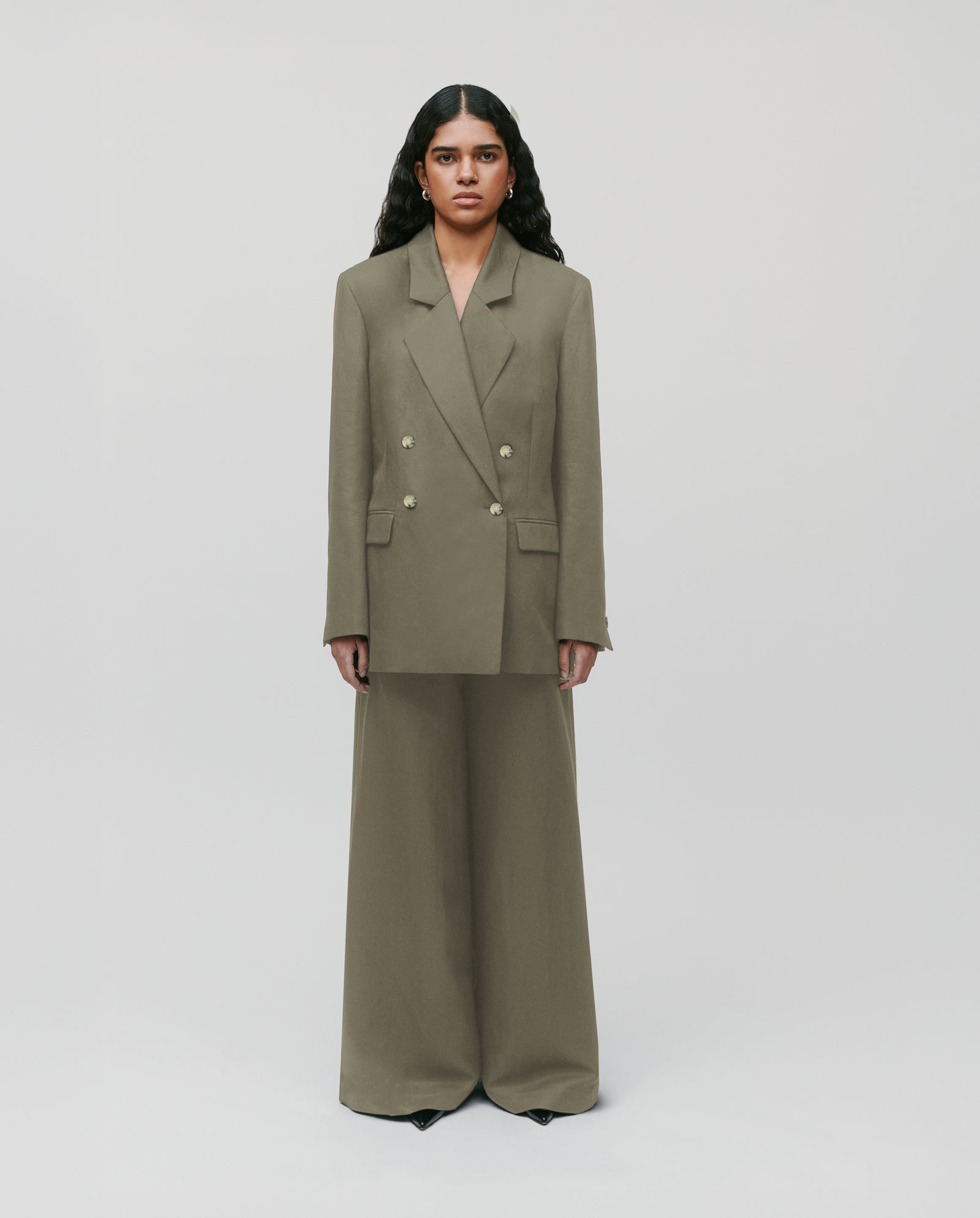 A woman stands facing forward, wearing an oversized olive green suit with wide-leg pants and a blazer.