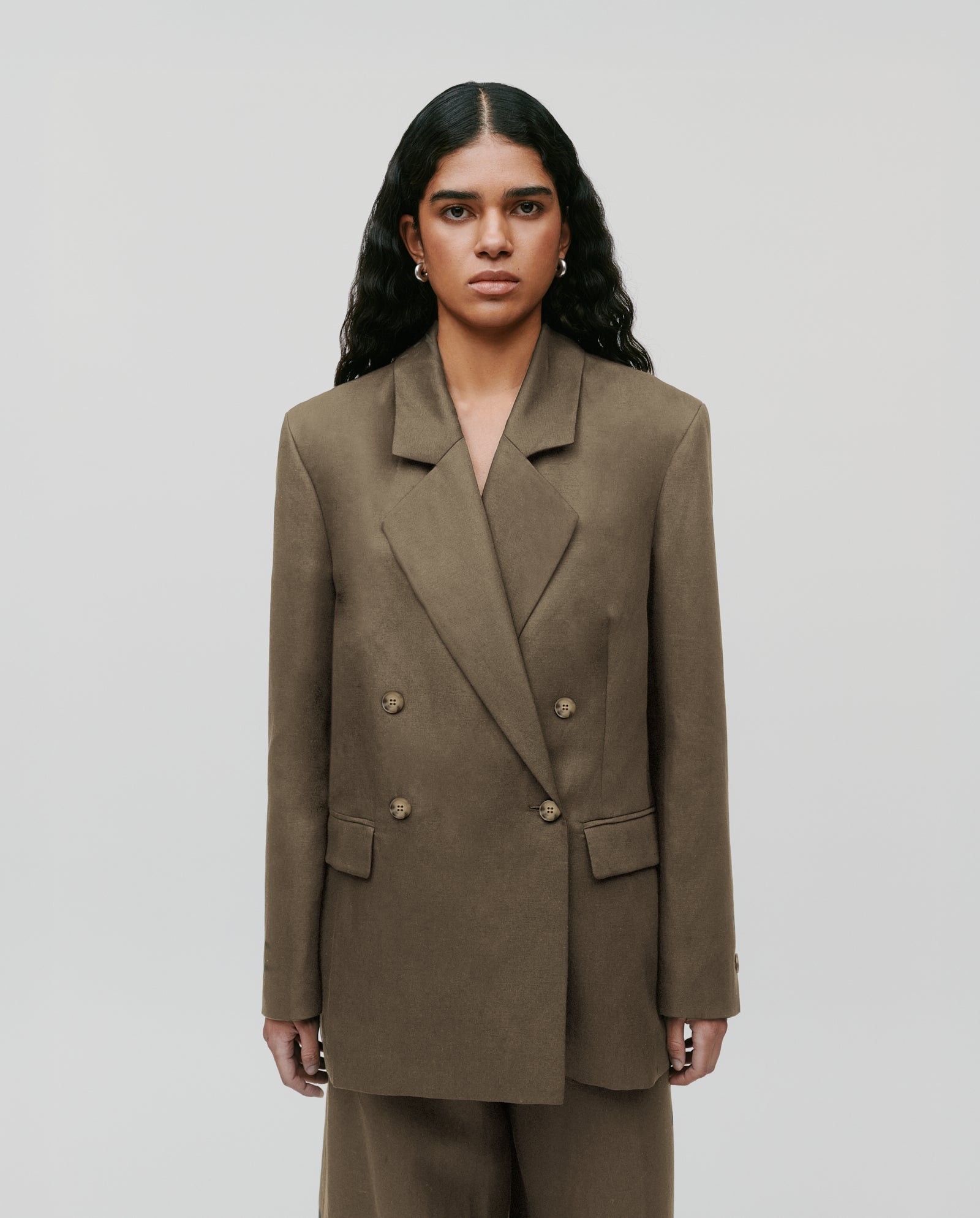 Woman with long wavy hair wearing the IVY OAK JASEMINA ROSE blazer and matching light linen pants in brown, standing against a plain background.