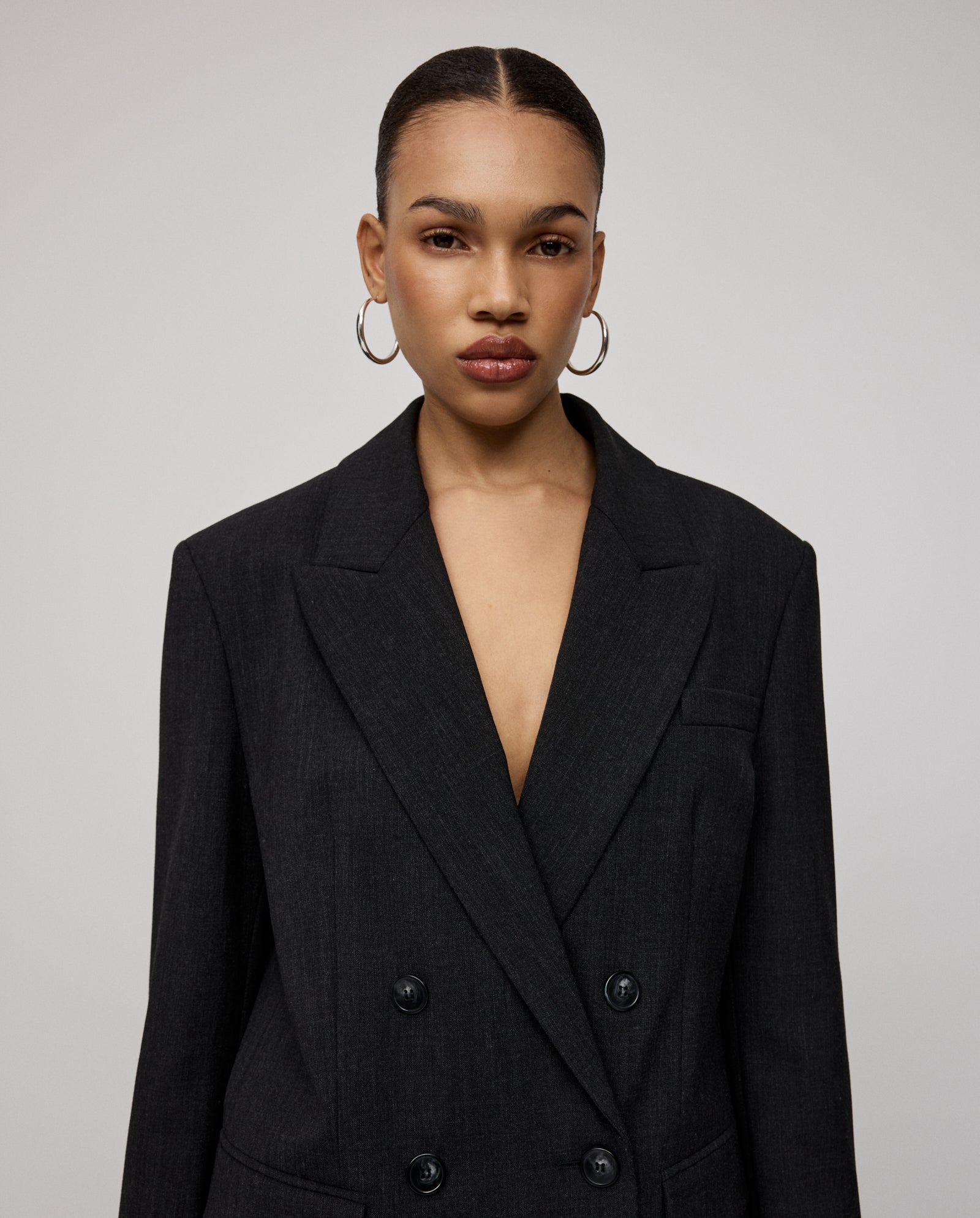 Woman with slicked-back hair wears hoop earrings and the IVY OAK JUDITH Blazer, a dark, double-breasted relaxed-fit jacket in wool-blend twill, set against a plain background.