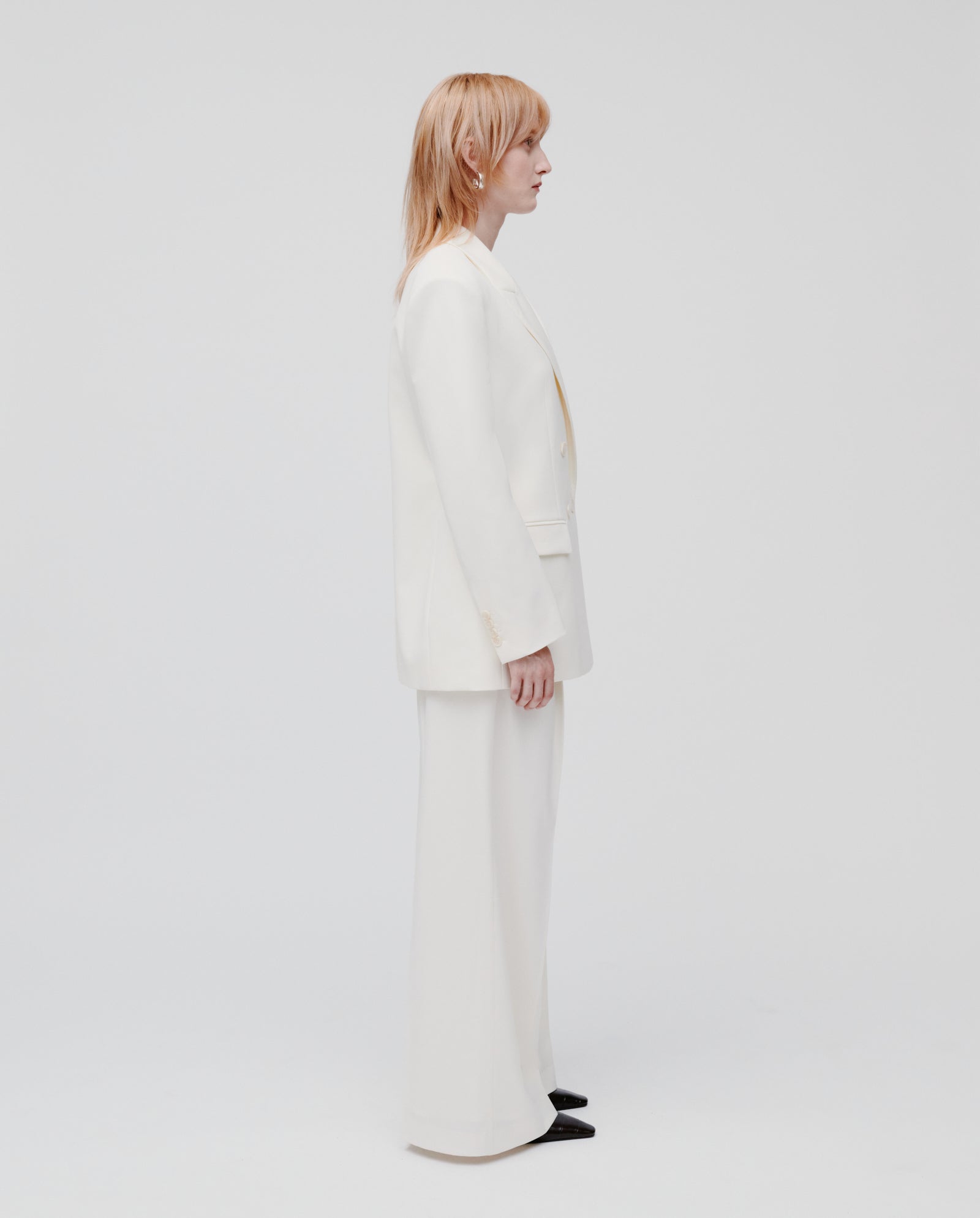 A person with straight light hair wears the IVY OAK JUDITH Blazer, a white lightweight wool-blend double-breasted suit, standing in profile against a plain white background.