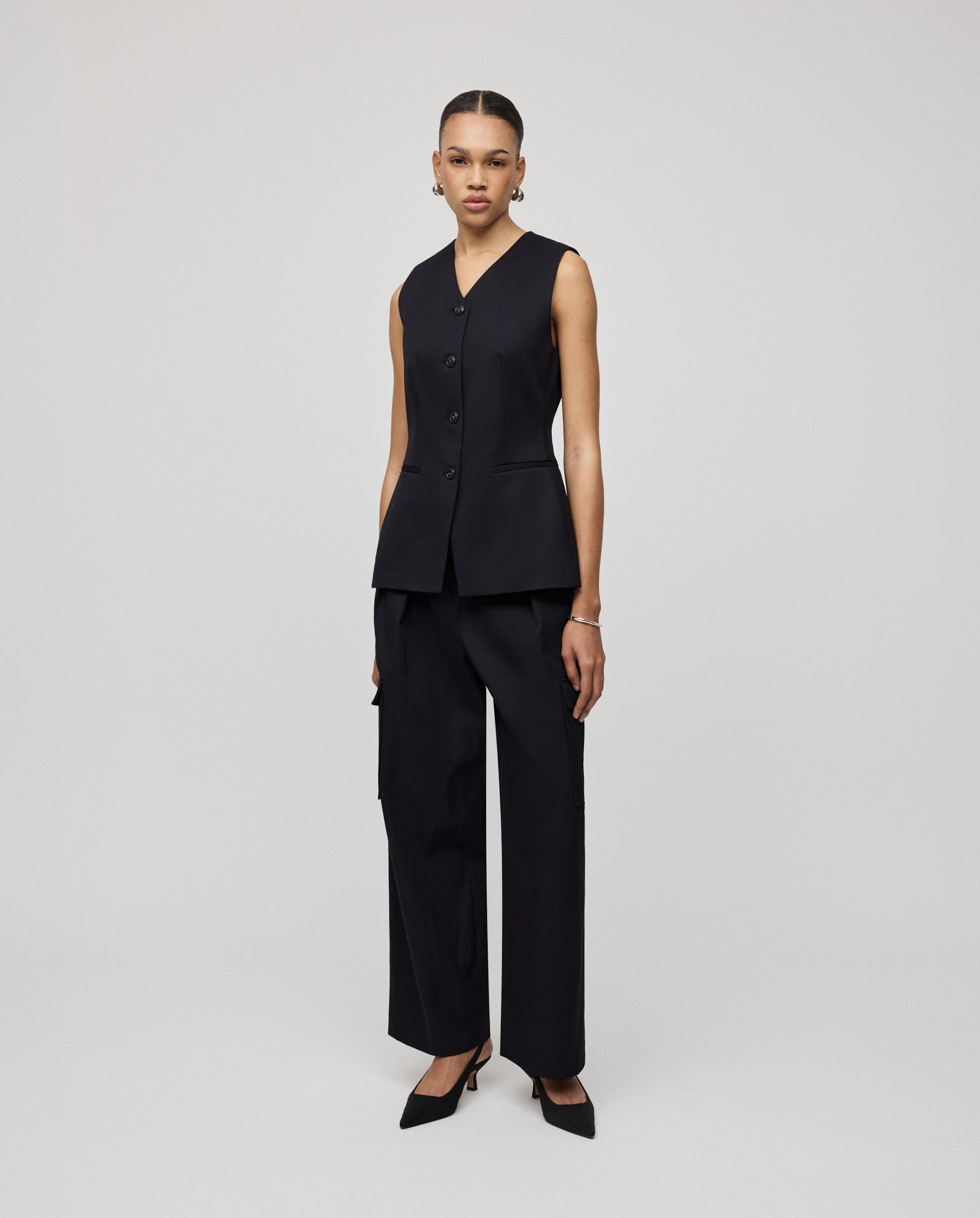 A woman wears the IVY OAK JUNIPER ROSE Vest, a sleeveless black blazer with cut-out details, paired with wide-leg black pants, highlighting her elegant silhouette against a plain background.