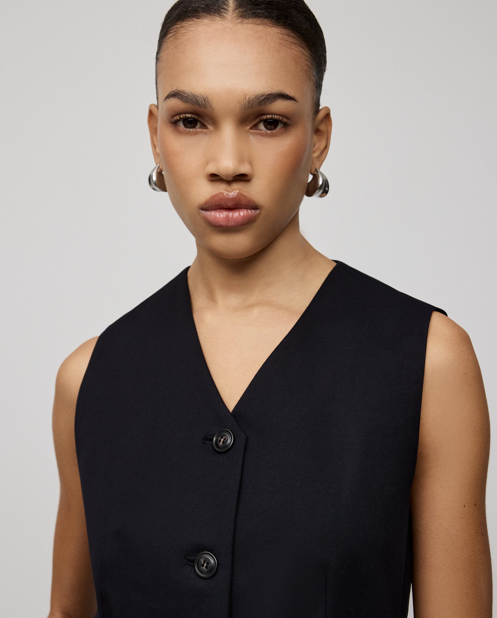 A woman with slicked-back hair wears the IVY OAK JUNIPER ROSE Vest—sleeveless, black, with cut-out detail—and hoop earrings, looking at the camera.