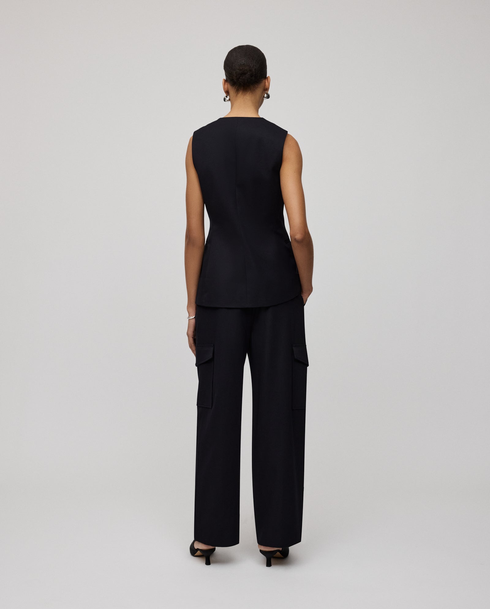 Wearing the IVY OAK JUNIPER ROSE Vest, a person stands with their back to the camera in black wide-leg pants, highlighting a striking sand glass silhouette.