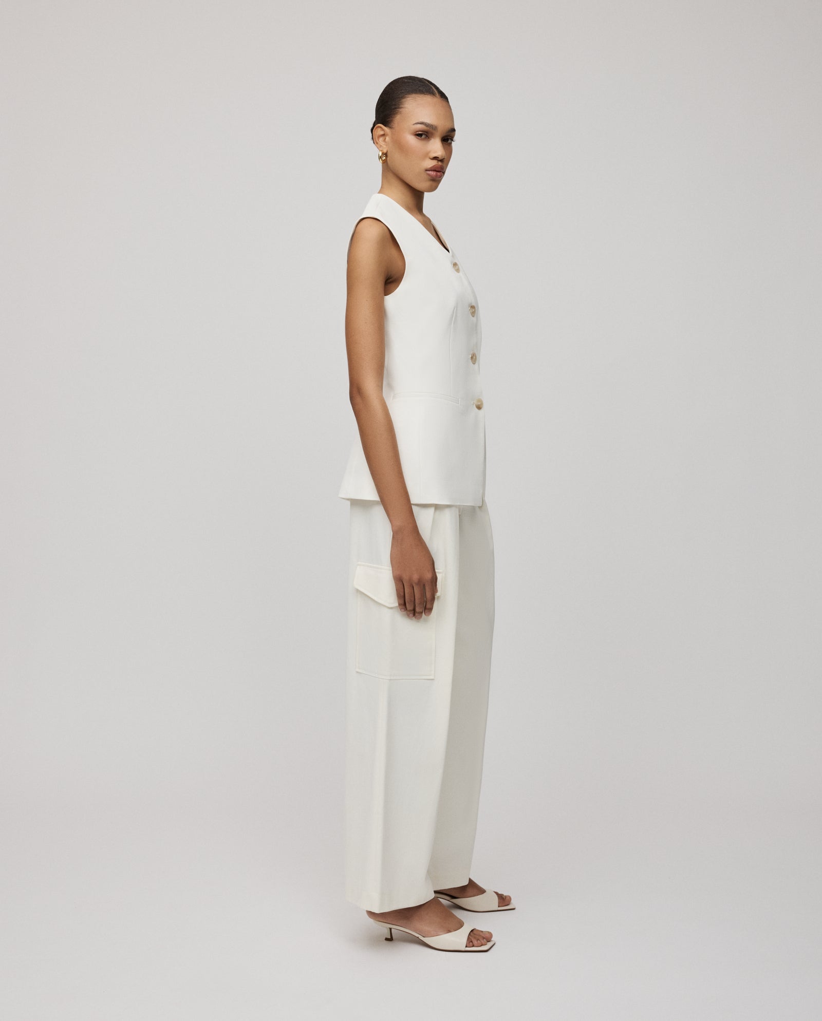 Woman wearing the IVY OAK JUNIPER ROSE Vest, a sleeveless white top with cut-out details, paired with wide-leg pants, standing sideways against a plain background.