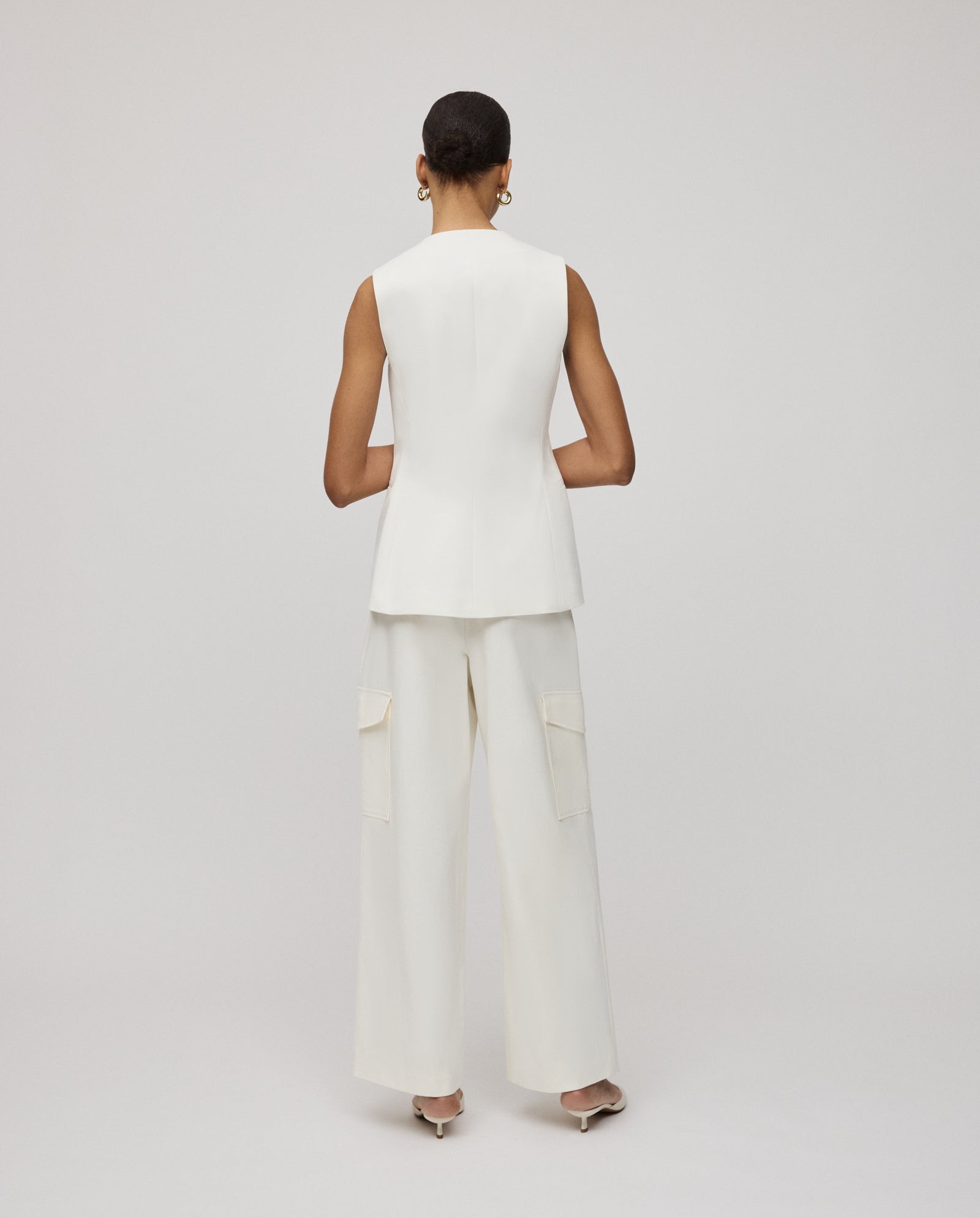 A woman stands with her back to the camera against a plain background, wearing the IVY OAK JUNIPER ROSE Vest—a sleeveless white top with button placket—paired with wide-leg pants.