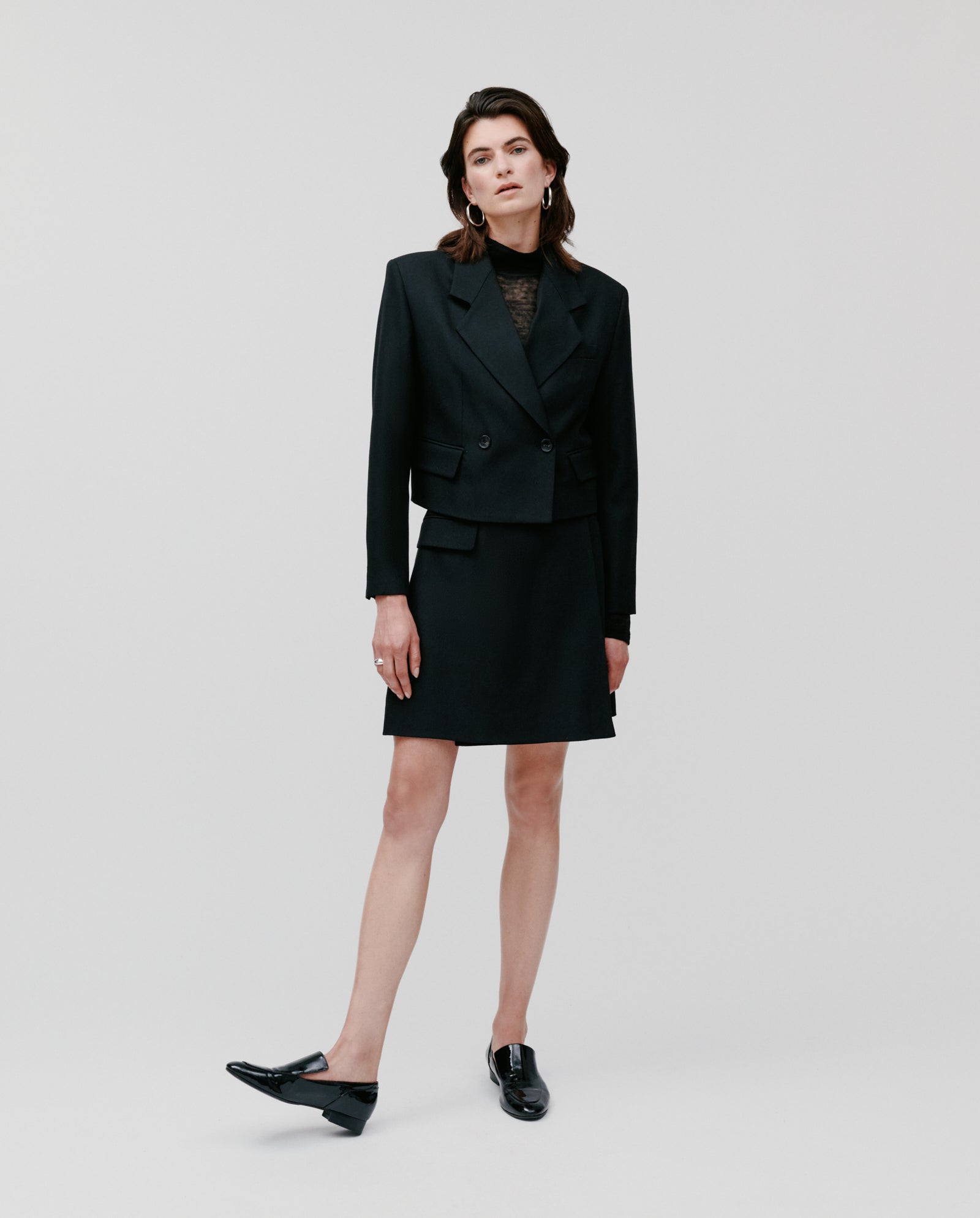 A woman wears the IVY OAK JENNY ANN Blazer, styled with a skirt, black loafers, and hoop earrings, standing against a plain white background and highlighting a modern, structured silhouette.
