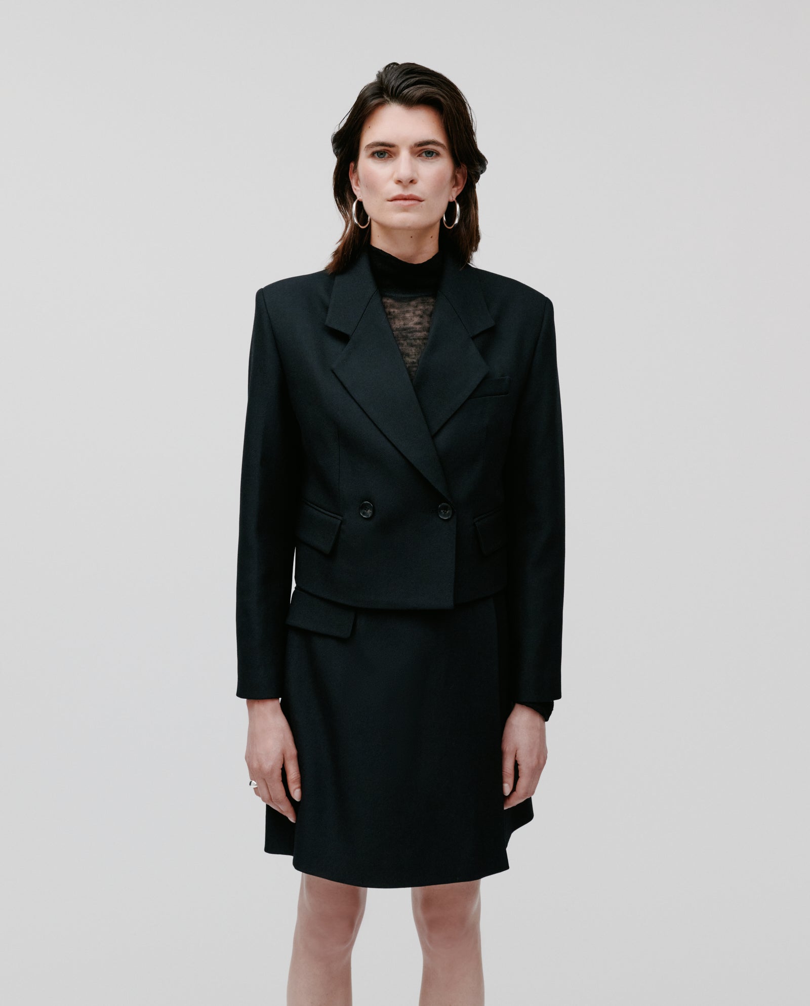 A woman models the IVY OAK JENNY ANN Blazer and skirt, styled with hoop earrings, standing against a plain white background to showcase the modern silhouette.