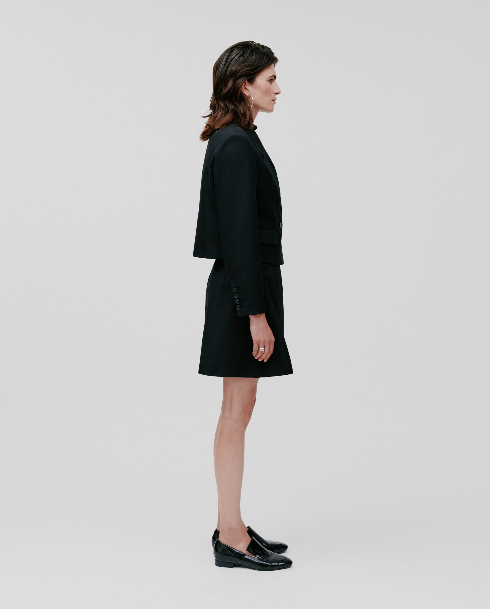 A woman stands in profile against a white background, showcasing the modern silhouette and structured tailoring of the IVY OAK JENNY ANN Blazer, styled with a skirt and loafers.