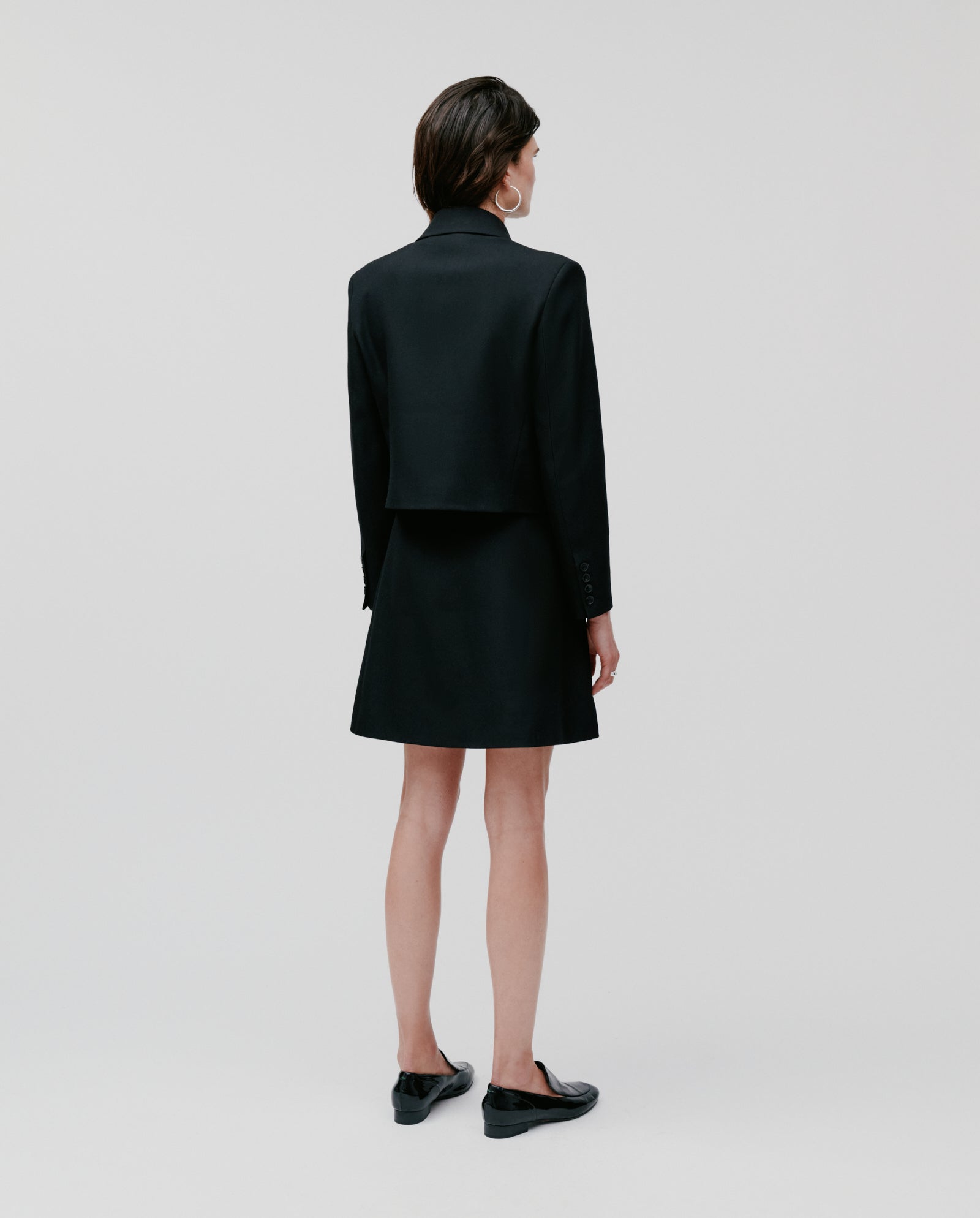 A person with short hair models the IVY OAK JENNY ANN Blazer and matching skirt, standing and facing away on a plain light background to highlight the blazer’s modern silhouette and structured tailoring.