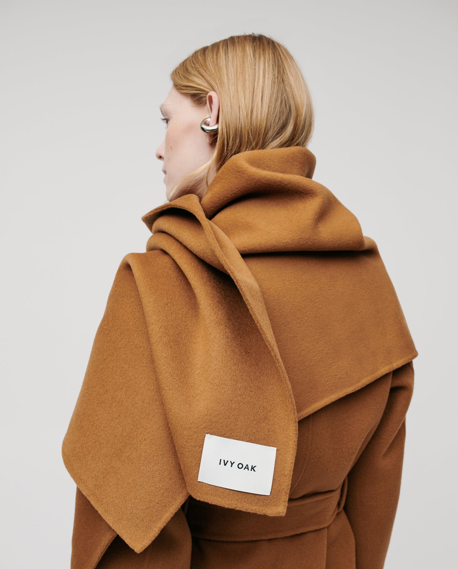 A person in a brown IVY OAK coat with a large collar, wearing an earbud, styles their look with the cozy AMY Scarf by IVY OAK for added warmth.